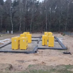 Wylany fundament pod dom, zabezpieczony czarną folią. Na fundamencie żółte bloczki Ytong. W tle las. Widoczne zbrojenia i łopata.
