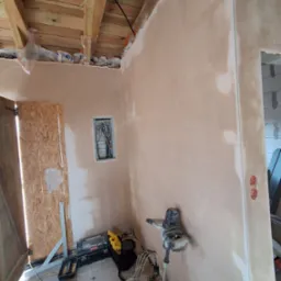 Ściana w trakcie gładzenia, widoczny mikser do gładzi, puszka elektryczna, drewniana konstrukcja dachu i narzędzia na podłodze. Prace wykończeniowe wewnątrz budynku.