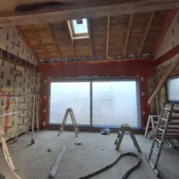 Wnętrze w trakcie budowy: ściany z pustaków, okno, drewniany strop z oknem dachowym, rusztowania i drabiny. Widoczny etap murowania ścian i montażu okna.