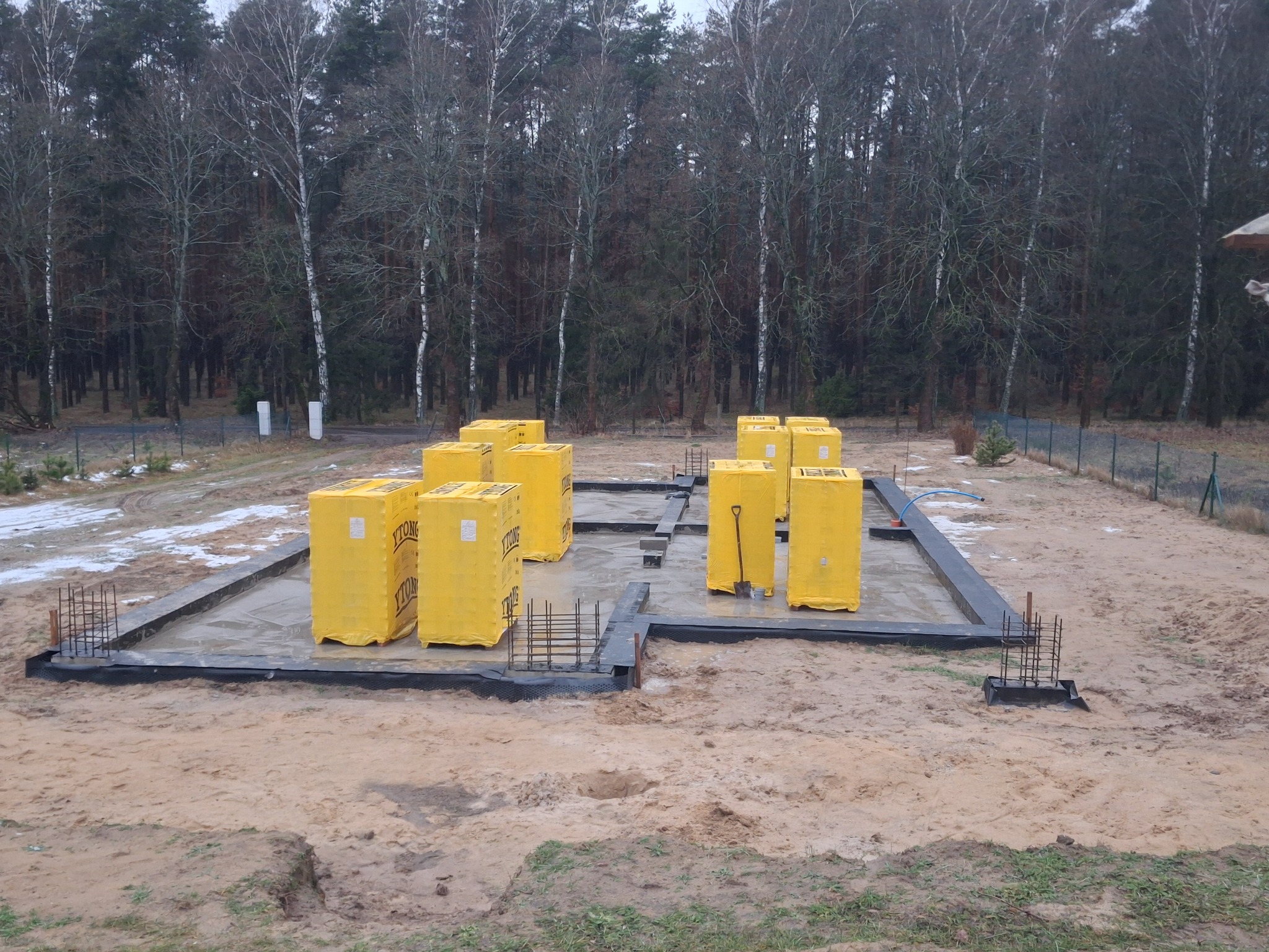 Wylany fundament pod dom, zabezpieczony czarną folią. Na fundamencie żółte bloczki Ytong. W tle las. Widoczne zbrojenia i łopata.
