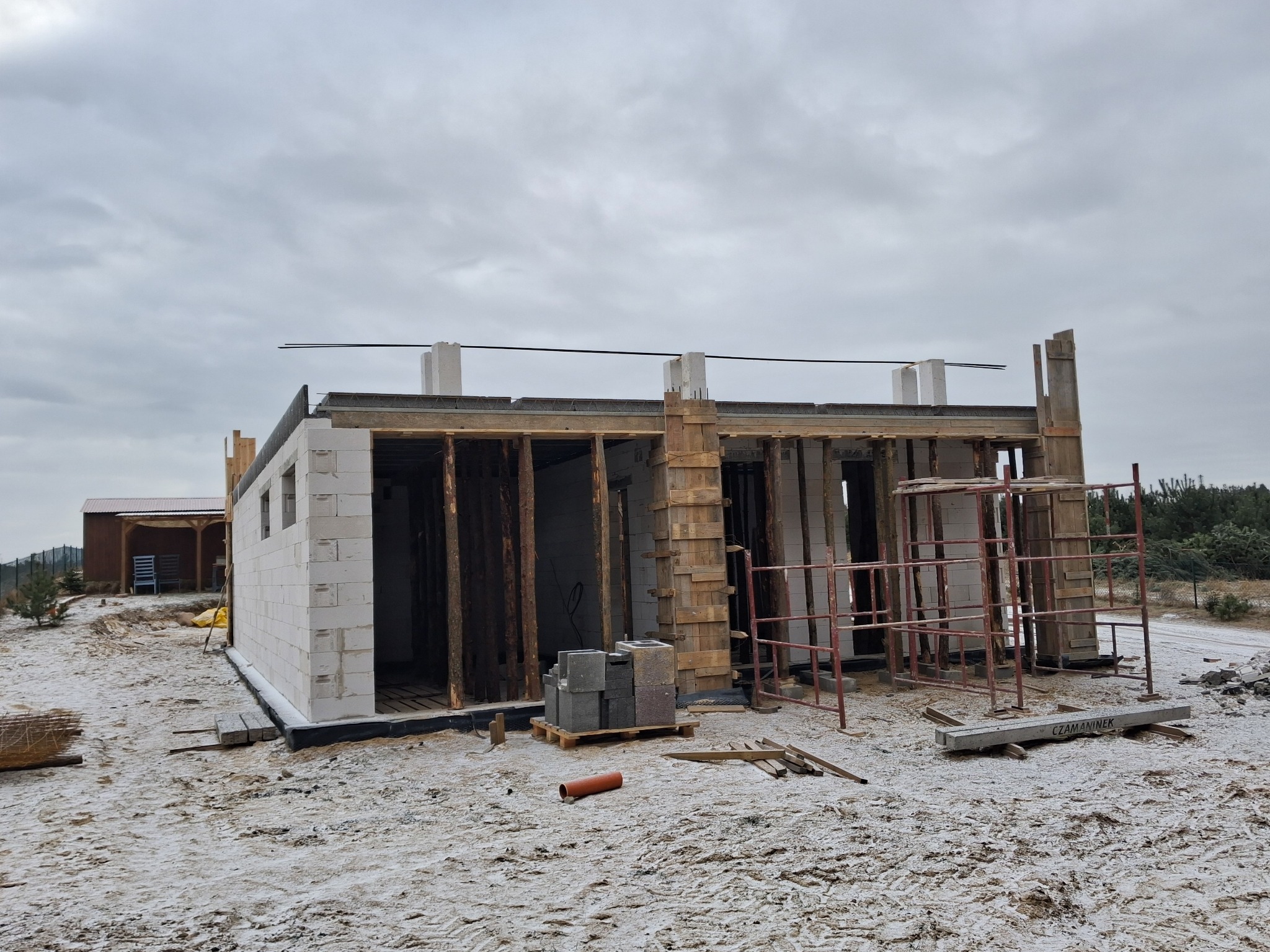 Niedokończony dom murowany z betonu komórkowego w zimowej scenerii. Widoczne drewniane szalunki, rusztowania i paleta z bloczkami.