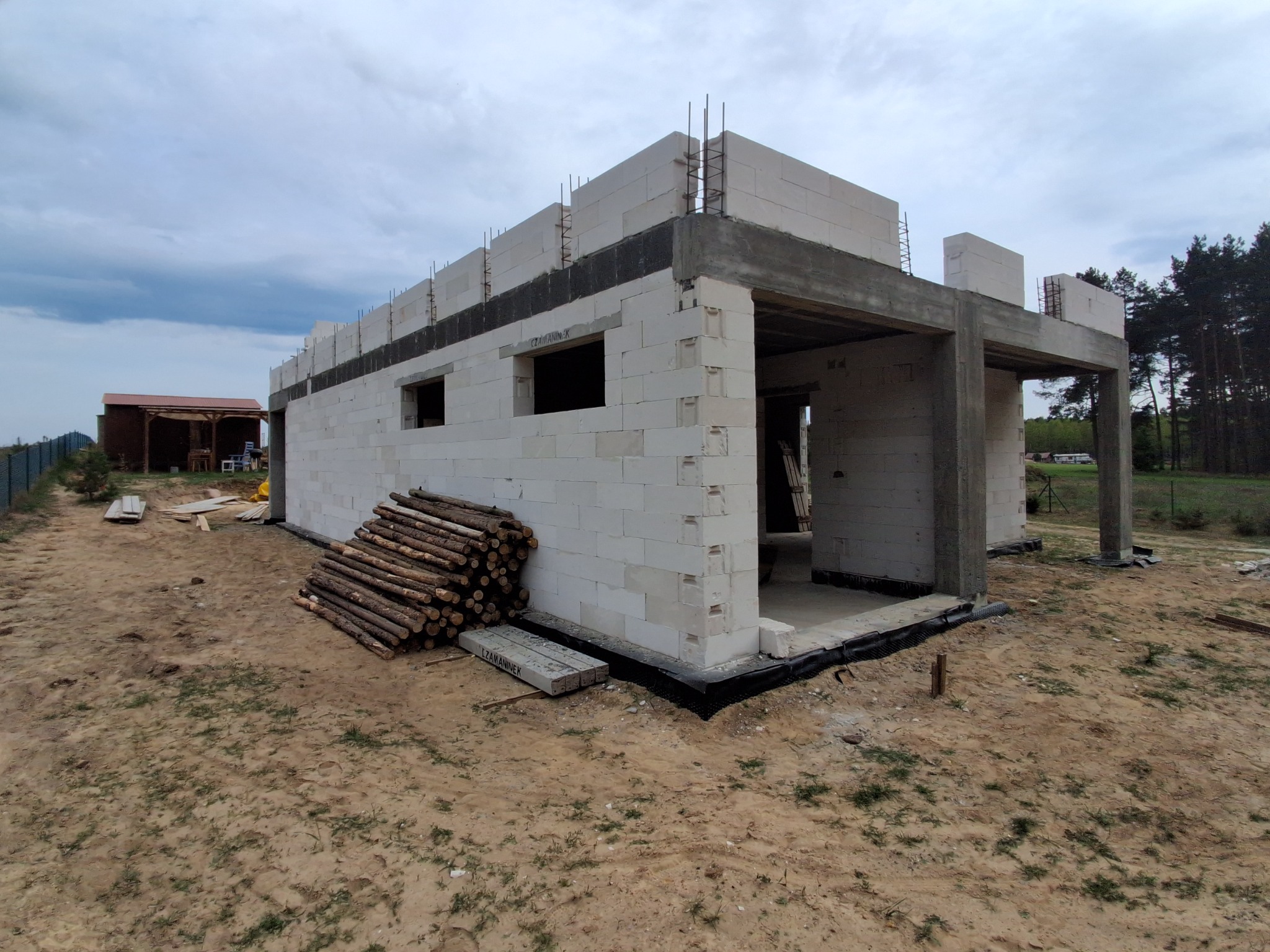 Surowy stan domu z białych bloczków, widoczne zbrojenia i betonowy wieniec. Na pierwszym planie stos drewna i materiały budowlane. W tle las i budynek gospodarczy.