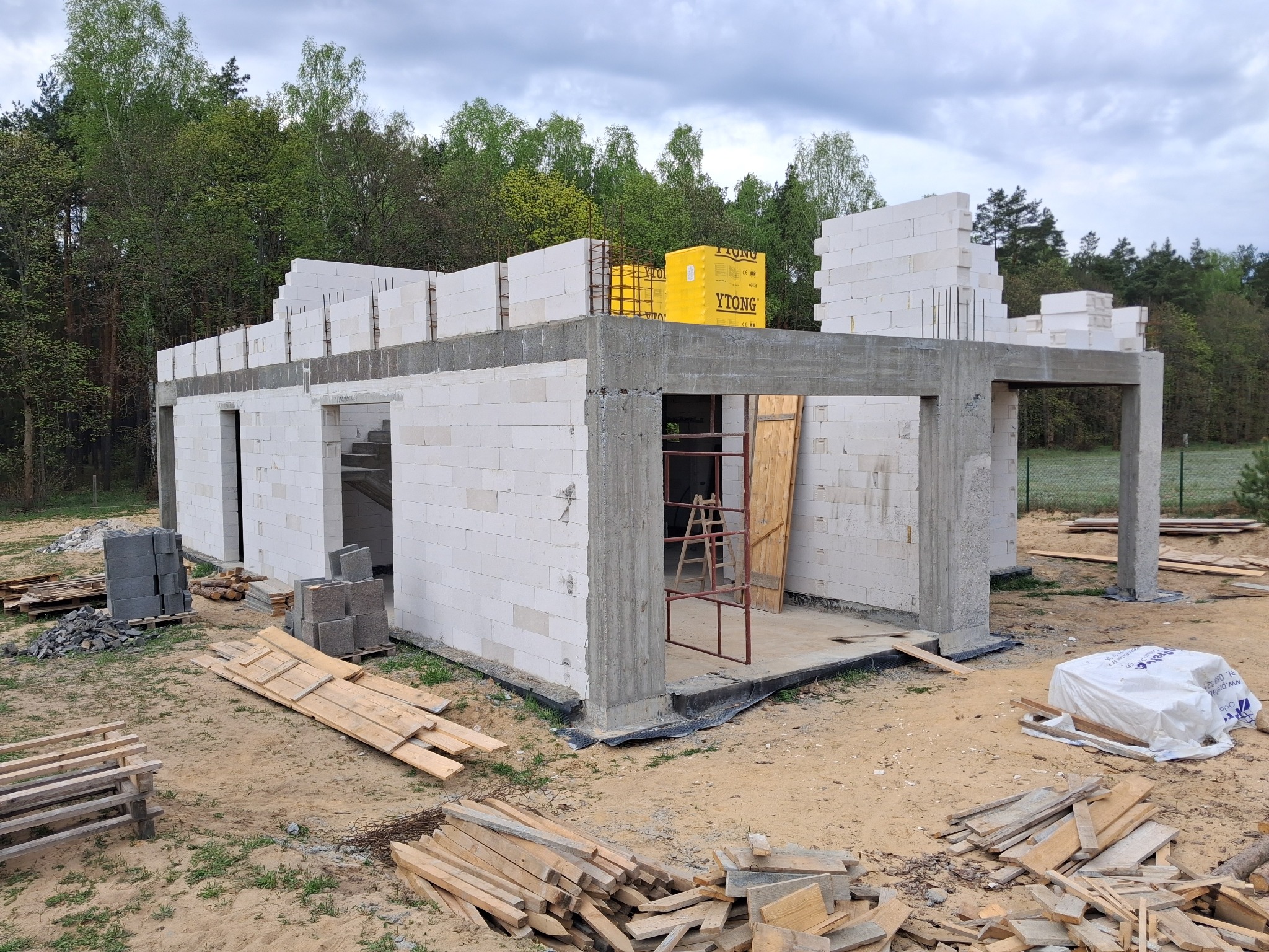 Budowa domu z betonu komórkowego Ytong w stanie surowym otwartym, widoczne zbrojenia, drewniane deski i rusztowanie, pakiet bloczków Ytong na stropie.