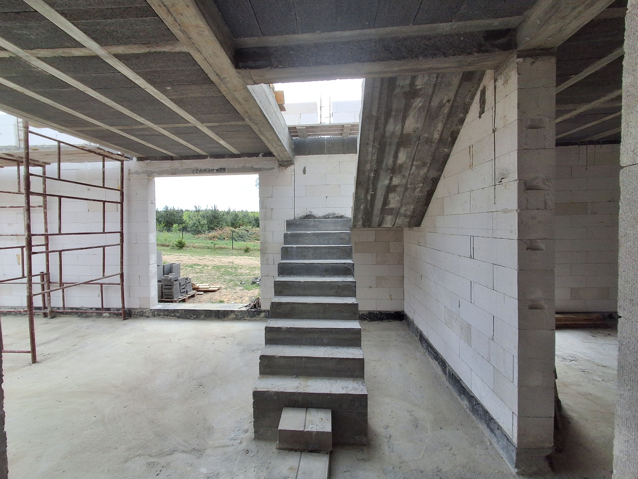 Surowe wnętrze domu w budowie: białe ściany z betonu komórkowego, betonowe schody i strop. Widoczne rusztowania i elementy konstrukcyjne w stanie surowym.