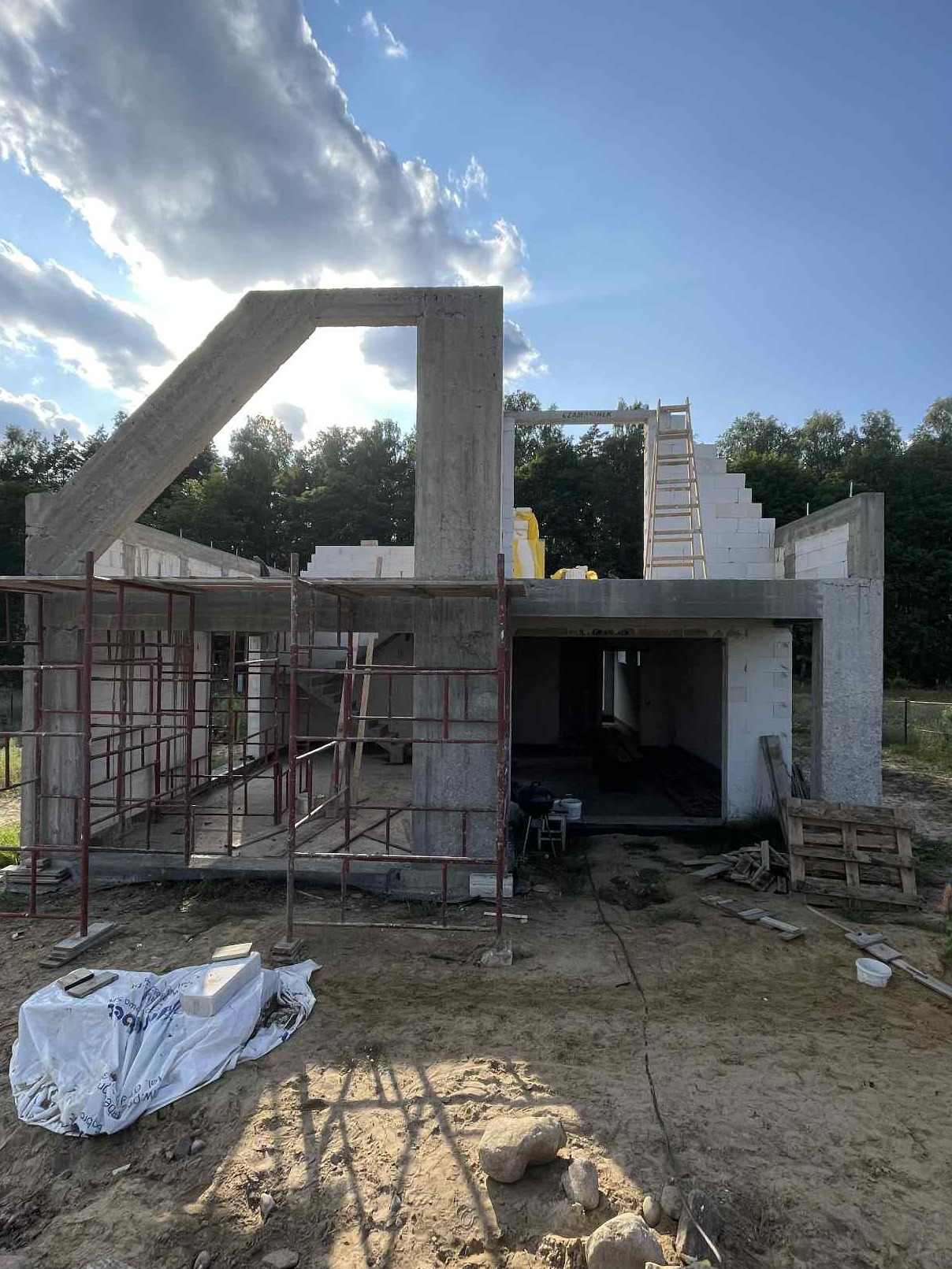 Surowy stan domu murowanego z betonu komórkowego w słoneczny dzień. Widoczne rusztowania, elementy konstrukcyjne i drewniana drabina.