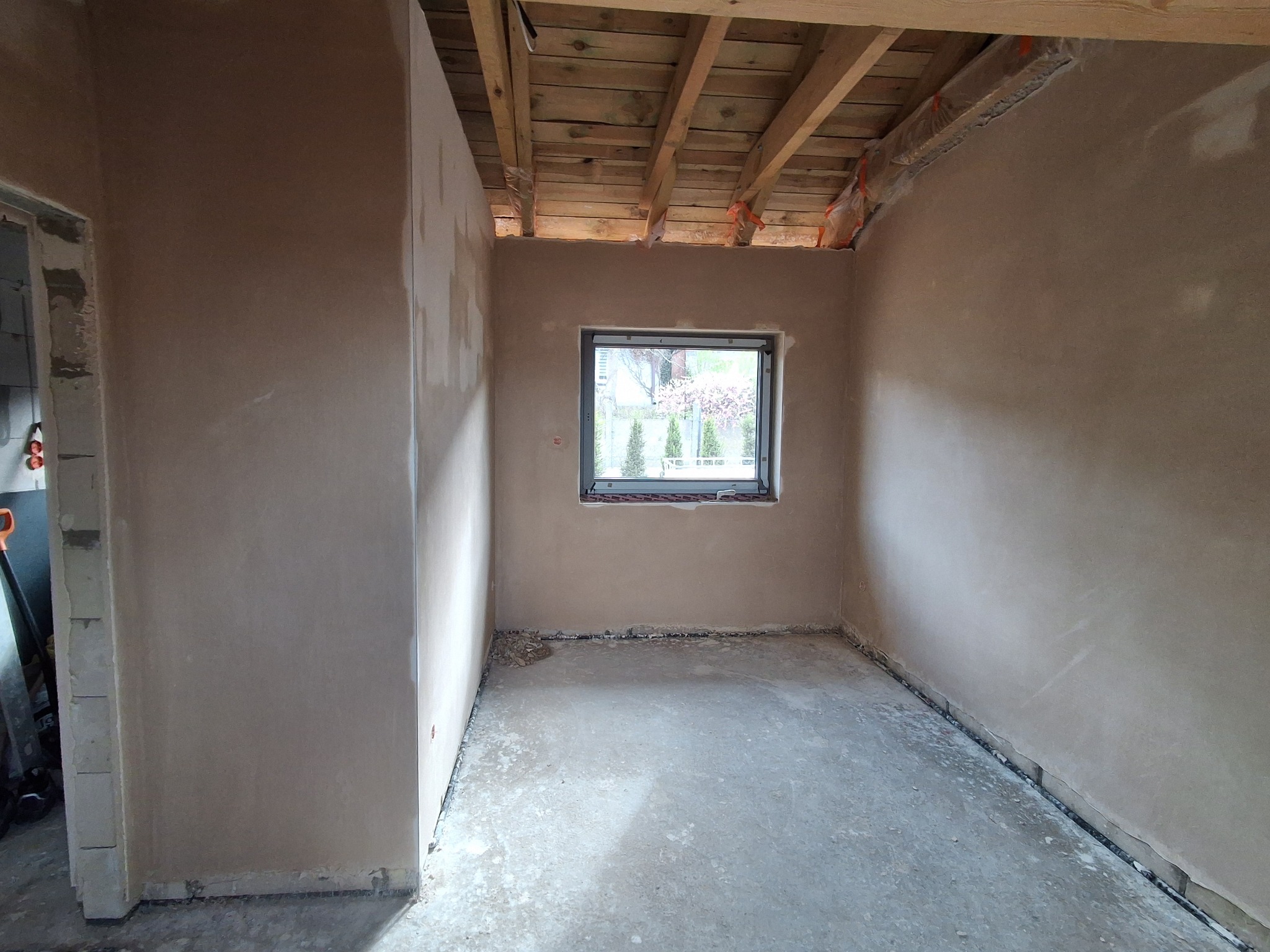 Wnętrze pokoju w stanie surowym: ściany pokryte jasnym tynkiem, drewniany strop, okno z widokiem na zieleń. Na podłodze wylewka. Widoczna surowa ściana z pustaków.