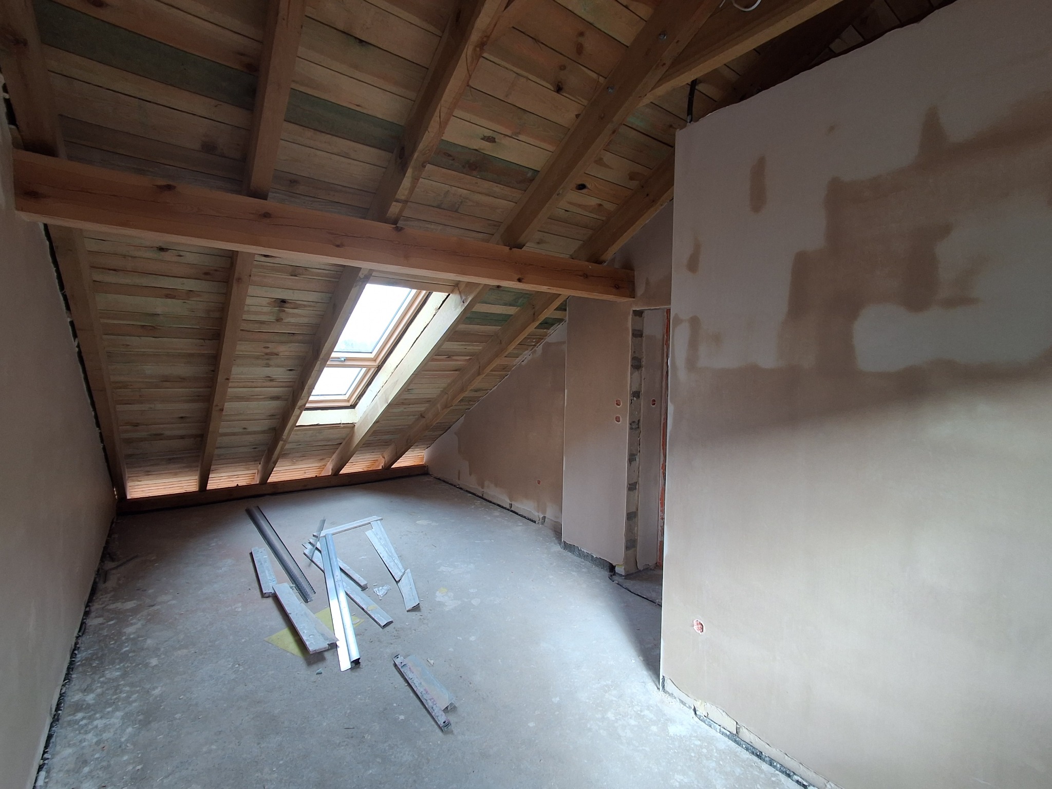 Poddasze w trakcie wykańczania: drewniana konstrukcja dachu z oknem, surowa podłoga i ściany w trakcie gładzenia, widoczne profile i listwy.