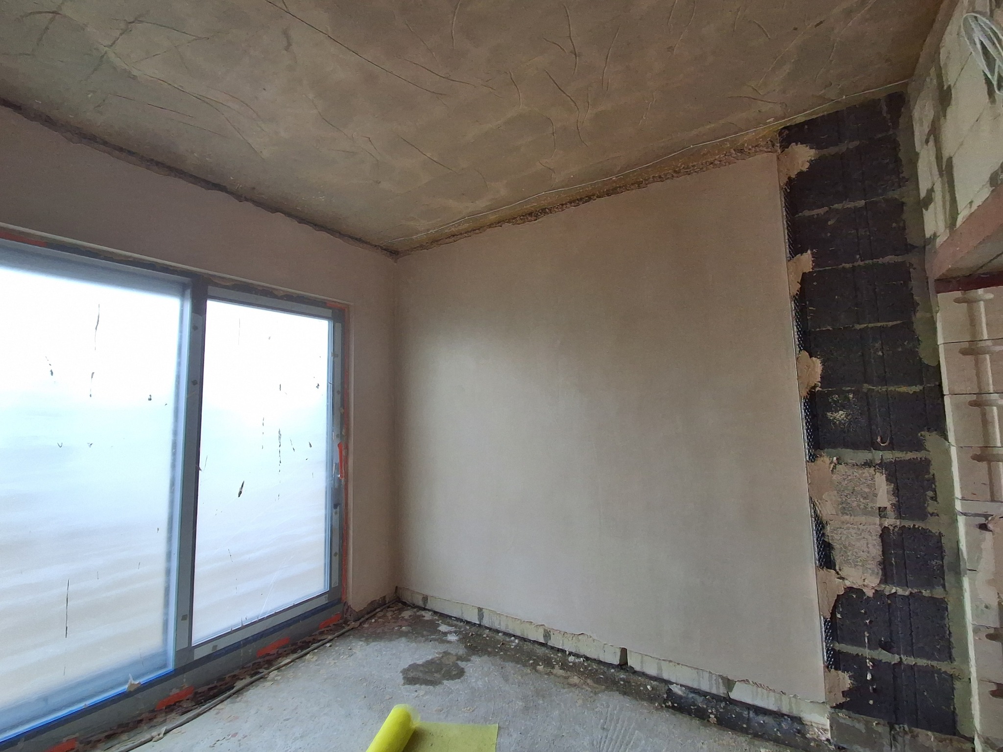 Wnętrze w trakcie wykończenia: gładzie na ścianach i suficie, okno z widokiem na zewnątrz, fragment ściany z widoczną strukturą bloczków. Na podłodze zwinięta mata.