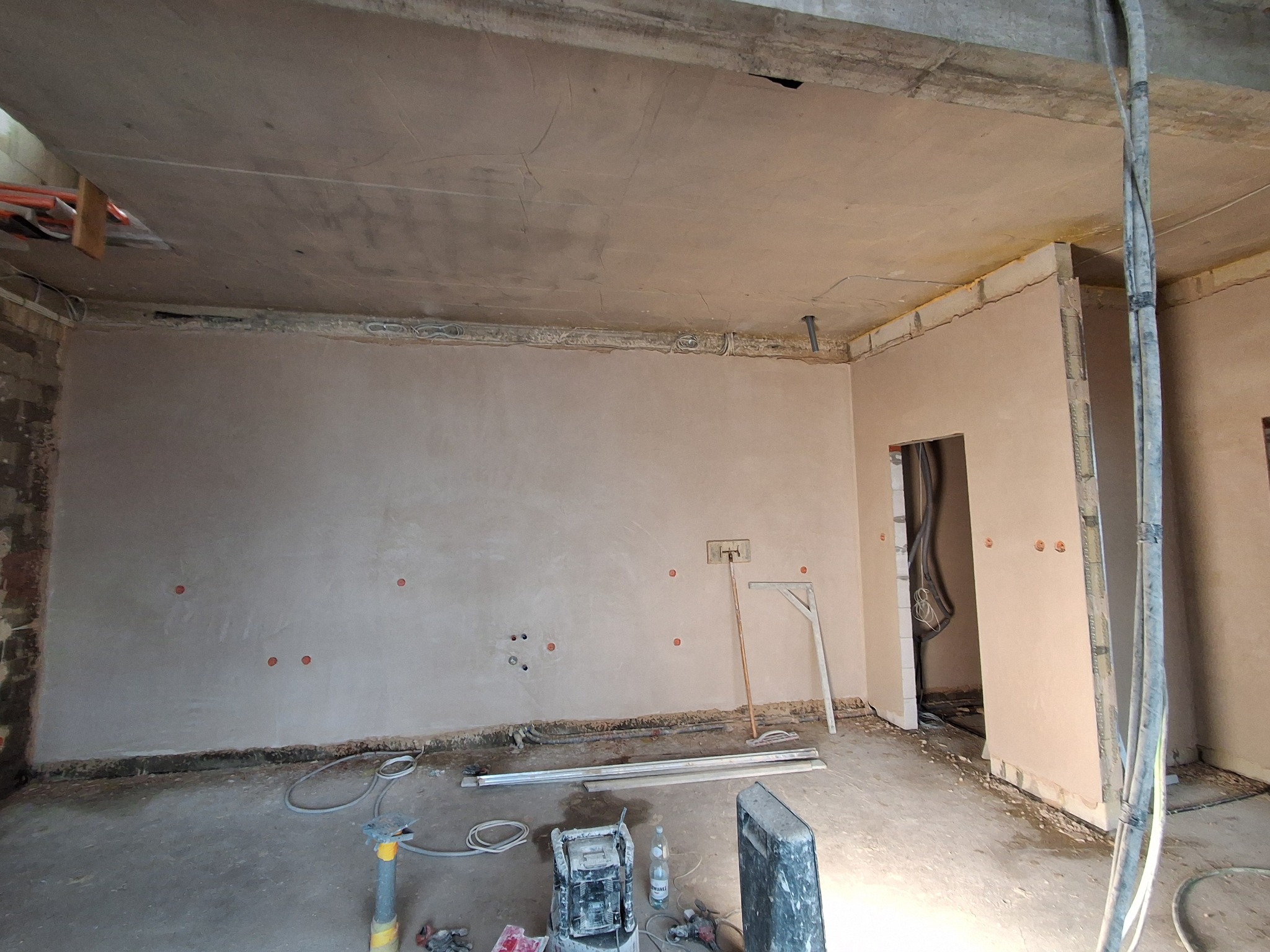 Wnętrze w trakcie prac wykończeniowych: ściany pokryte gładzią, widoczne instalacje elektryczne, narzędzia i materiały budowlane. Surowy stan pomieszczenia przed malowaniem.