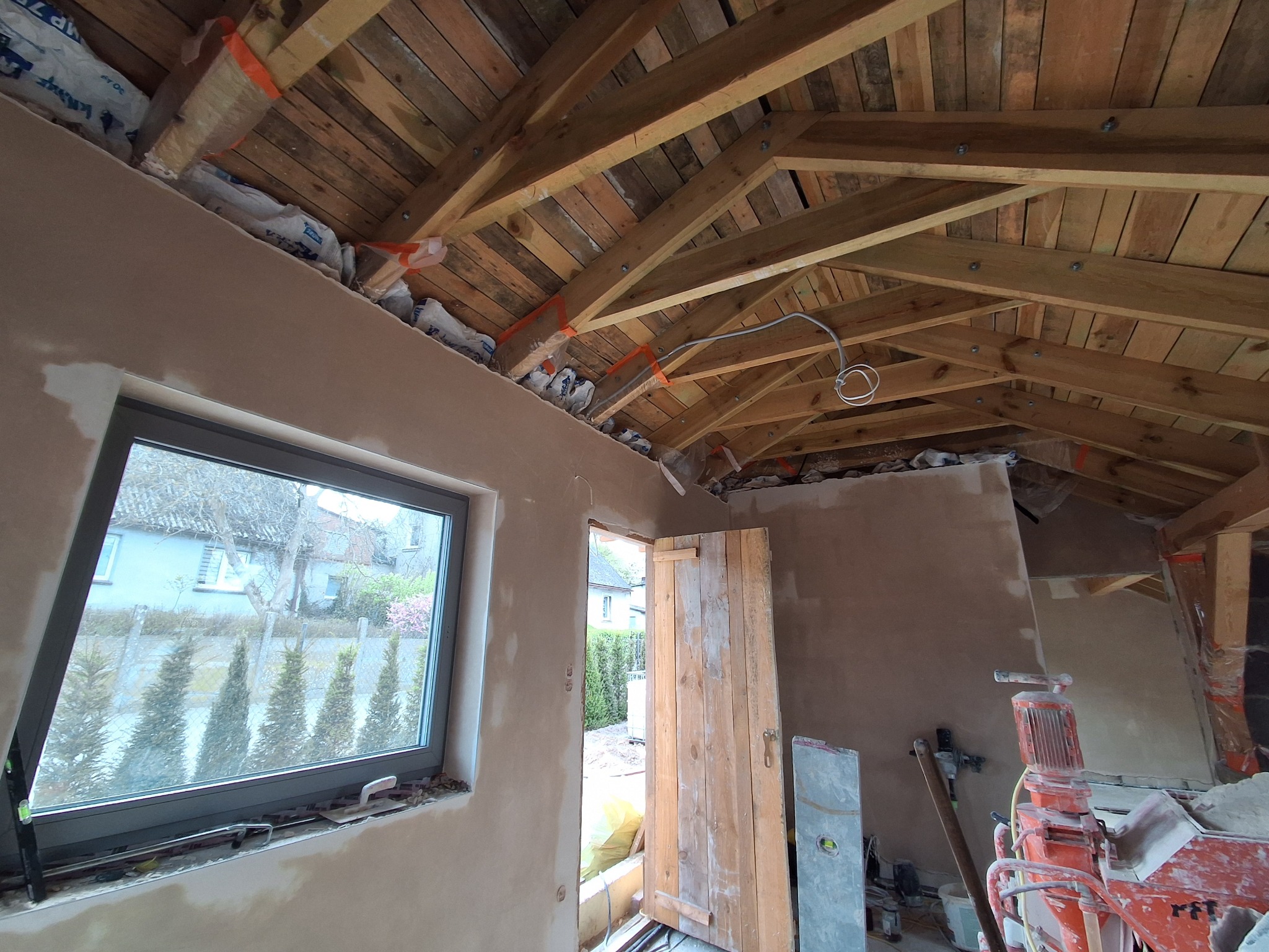 Wnętrze w trakcie prac wykończeniowych: drewniana więźba dachowa, okno z widokiem na zieleń, otynkowane ściany i narzędzia budowlane. Widoczna izolacja dachu.