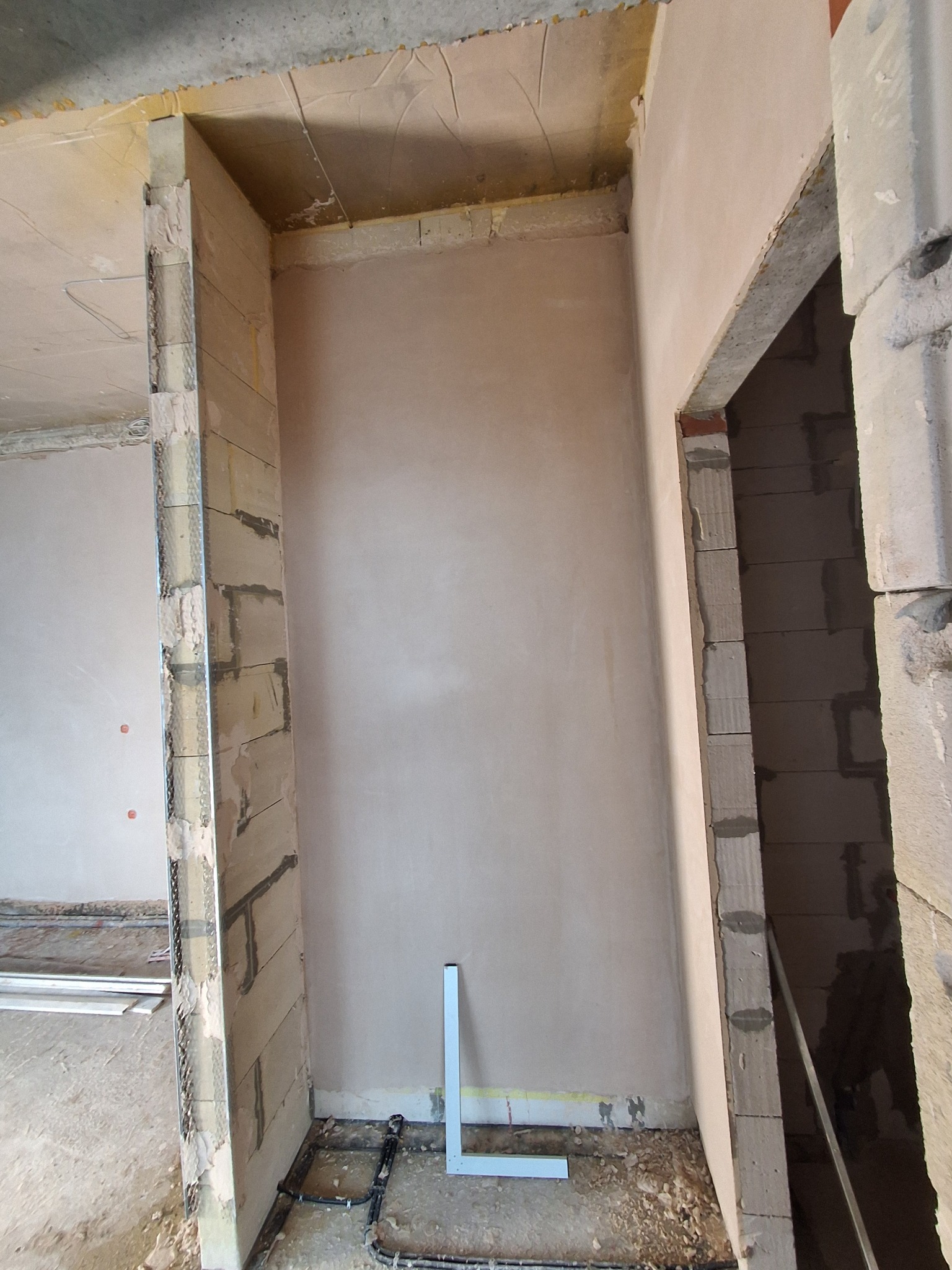 Wnętrze w trakcie prac wykończeniowych: ściana po gładziowaniu, widoczne fragmenty konstrukcji z pustaków i metalowe profile. Na podłodze elementy instalacji.