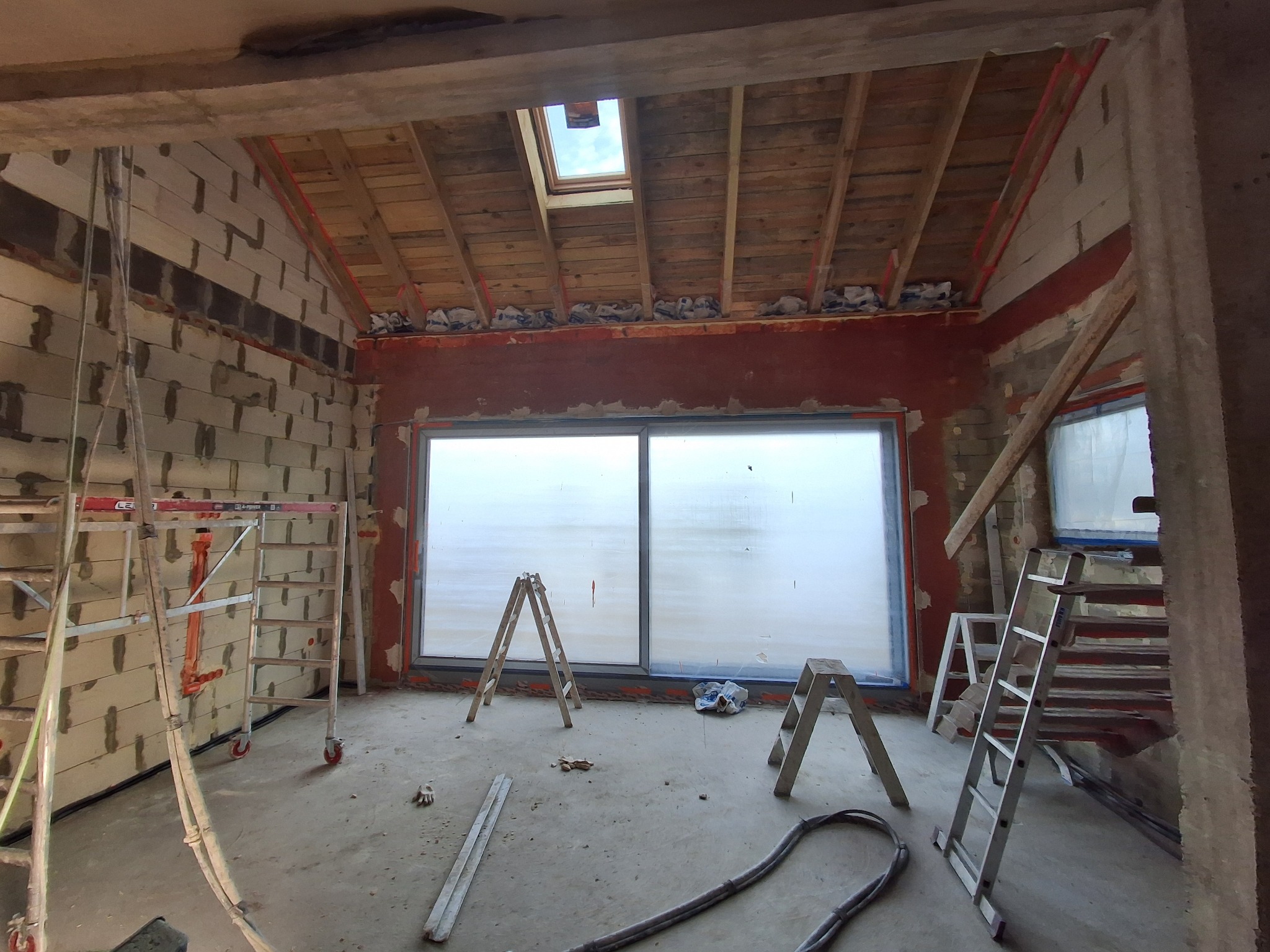 Wnętrze w trakcie budowy: ściany z pustaków, okno, drewniany strop z oknem dachowym, rusztowania i drabiny. Widoczny etap murowania ścian i montażu okna.