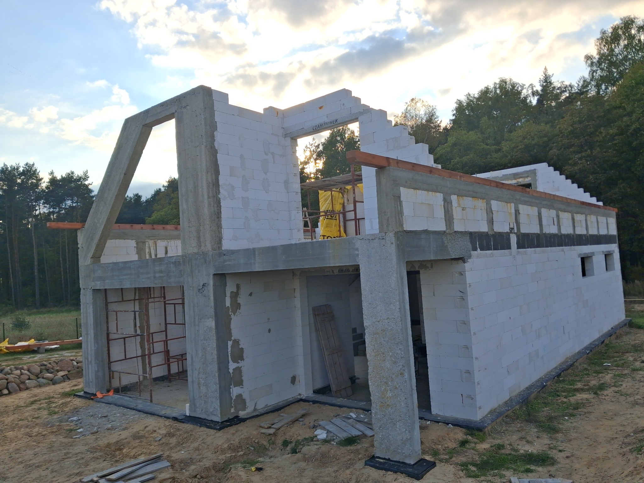 Surowy stan domu z betonu komórkowego w trakcie budowy, widoczne ściany, słupy i nadproża, zarys okien i drzwi, teren budowy z piaskiem i drewnianymi elementami.