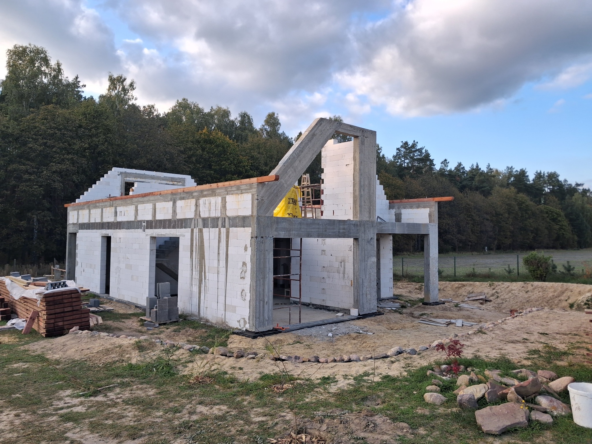 Budowa domu jednorodzinnego w trakcie wznoszenia ścian z betonu komórkowego. Widoczne elementy konstrukcyjne i materiały budowlane na tle zieleni i błękitnego nieba.