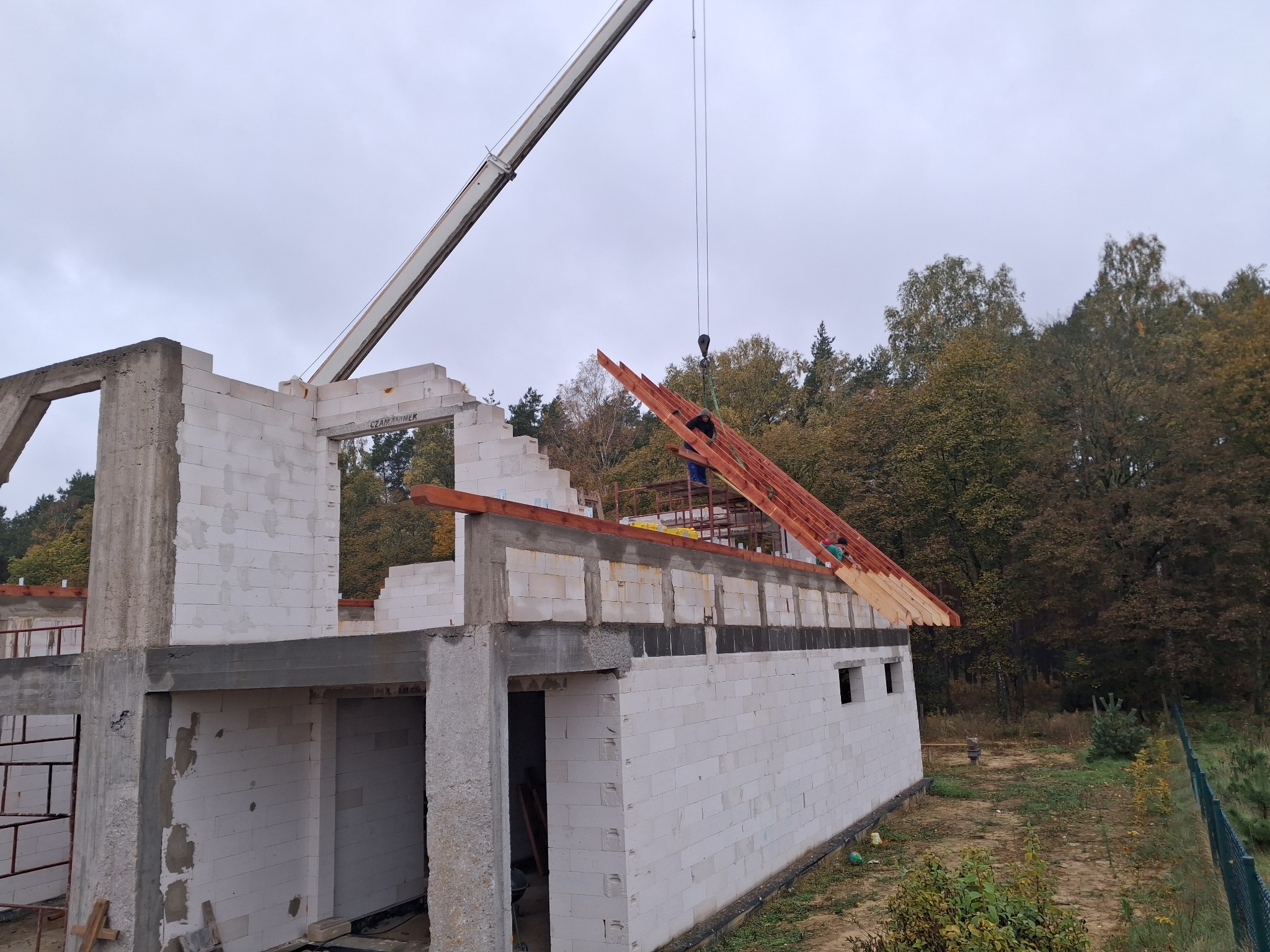 Wznoszenie domu z białych bloczków: dźwig podnosi elementy więźby dachowej. Widoczne zbrojenia i betonowe nadproża. W tle drzewa i pochmurne niebo.