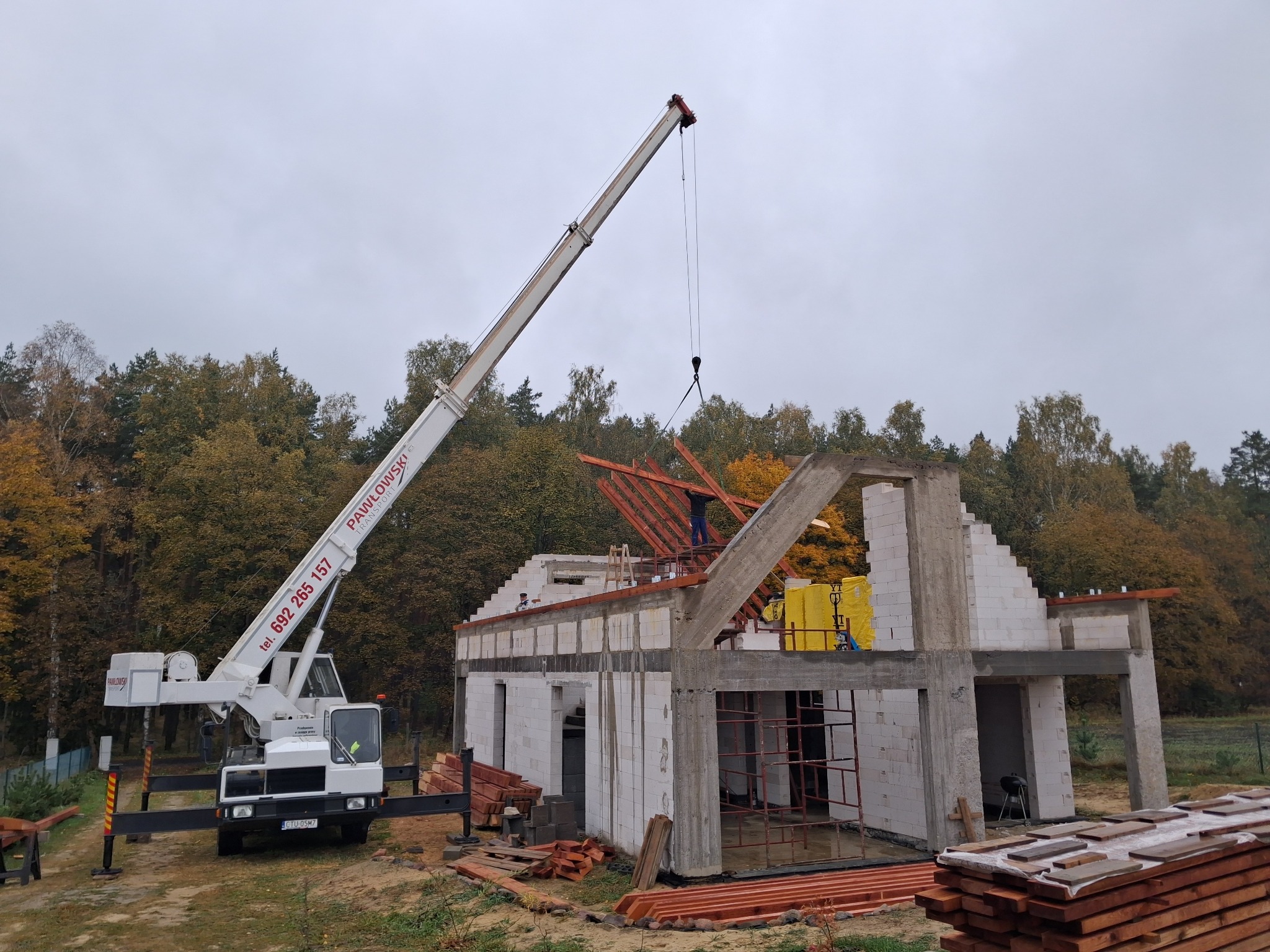 Budowa domu w stanie surowym otwartym z użyciem dźwigu. Widoczne ściany z betonu komórkowego, drewniana więźba dachowa i dźwig Pawłowski na tle lasu.