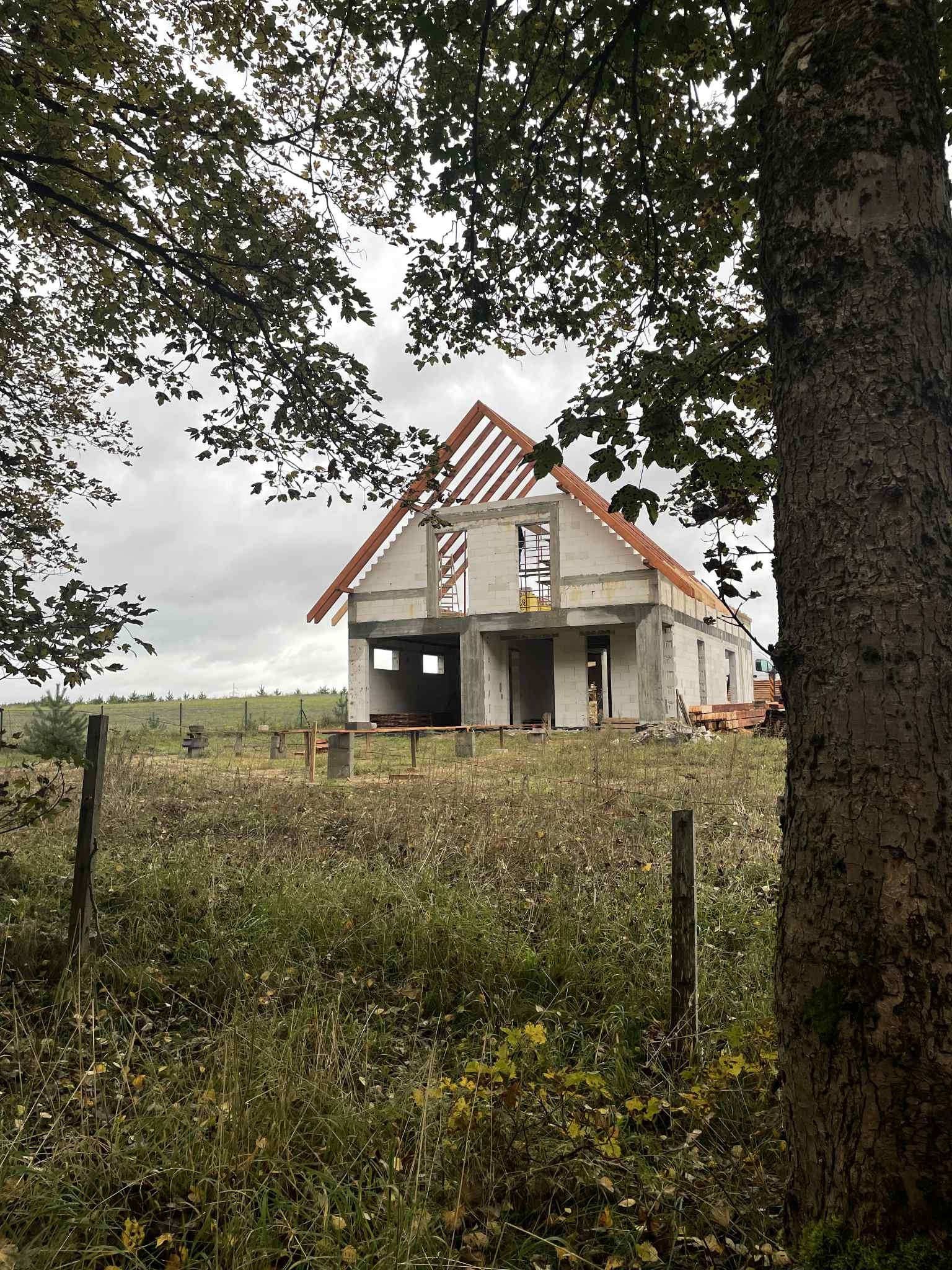 Surowy stan domu z betonu komórkowego w trakcie budowy, widoczny przez drzewa. Dach w konstrukcji drewnianej, otoczenie naturalne, jesienne.