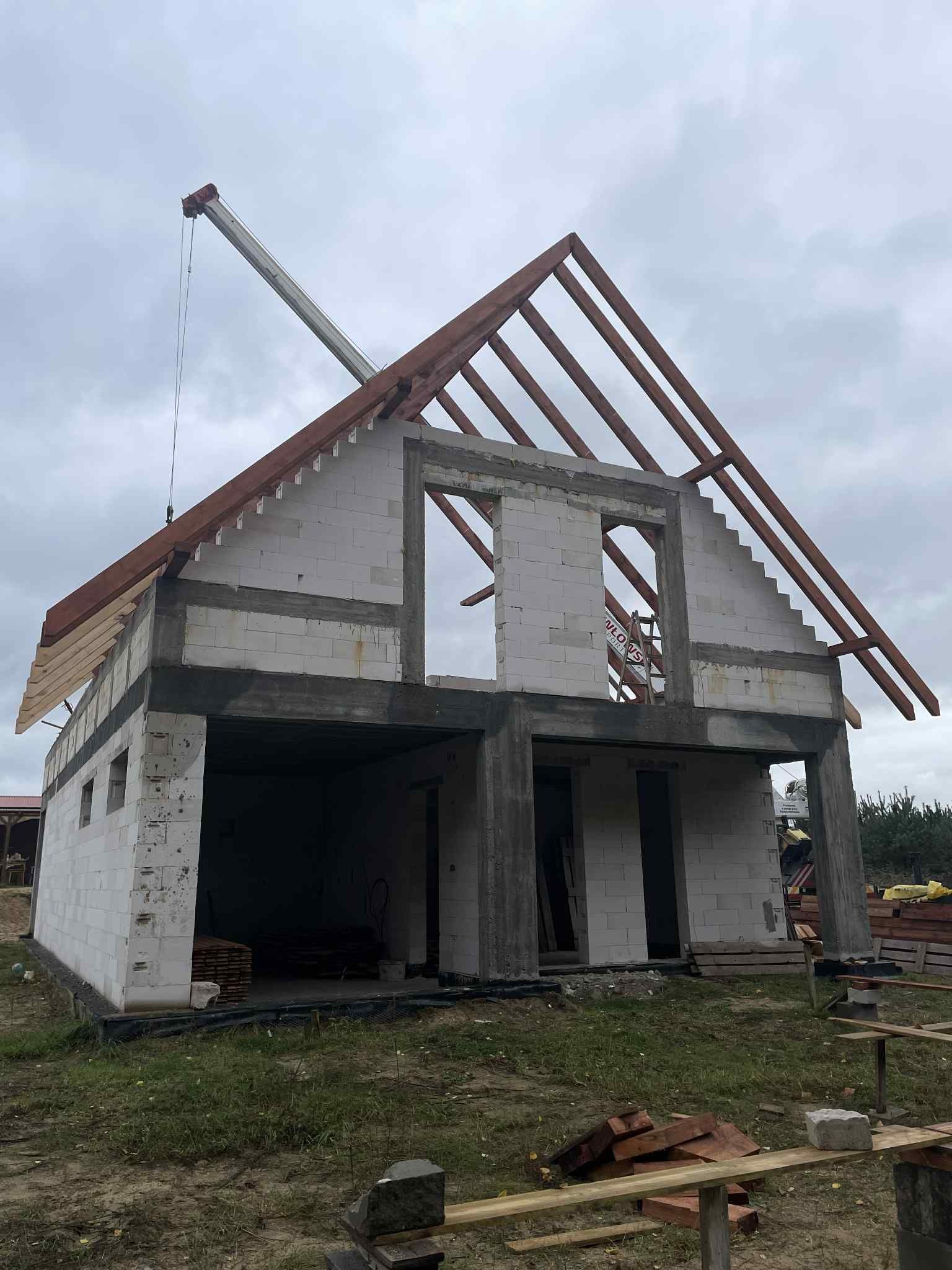 Surowy stan domu z białego bloczka, z widoczną konstrukcją dachu i dźwigiem w tle, w pochmurny dzień. Widoczne otwory okienne i drzwiowe.