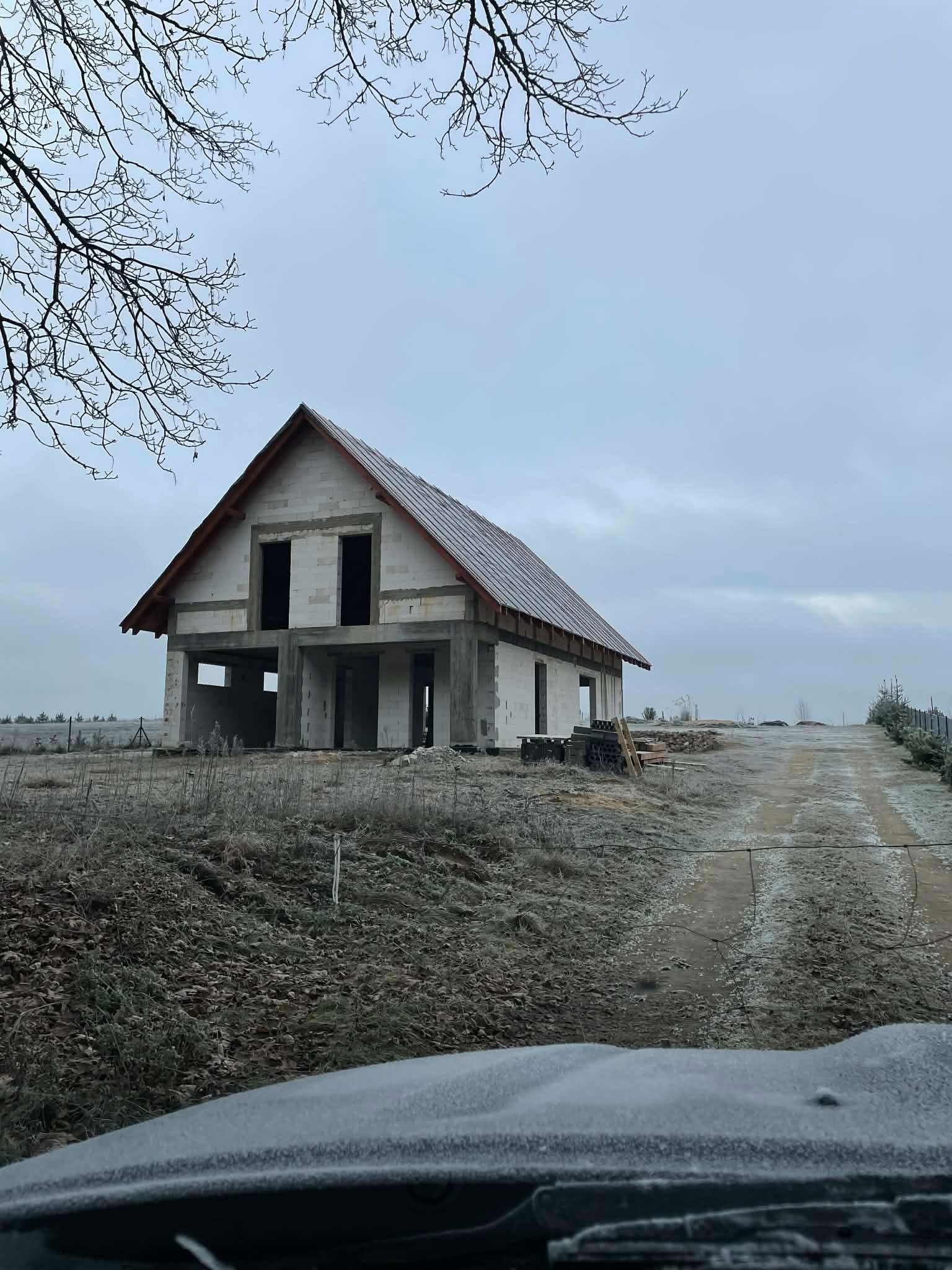 Surowy stan domu z białego pustaka, z częściowo ukończonym dachem, na tle pochmurnego nieba i zimowego krajobrazu. Widok z perspektywy kierowcy samochodu.