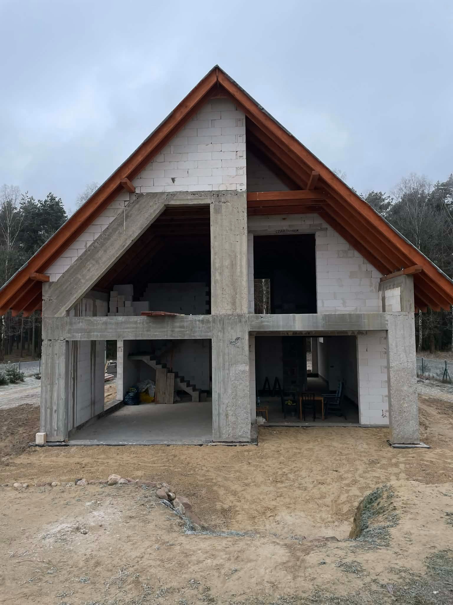 Surowy stan domu z betonu komórkowego i drewnianą więźbą dachową, widoczne schody wewnętrzne i meble wewnątrz, teren wokół w stanie naturalnym.