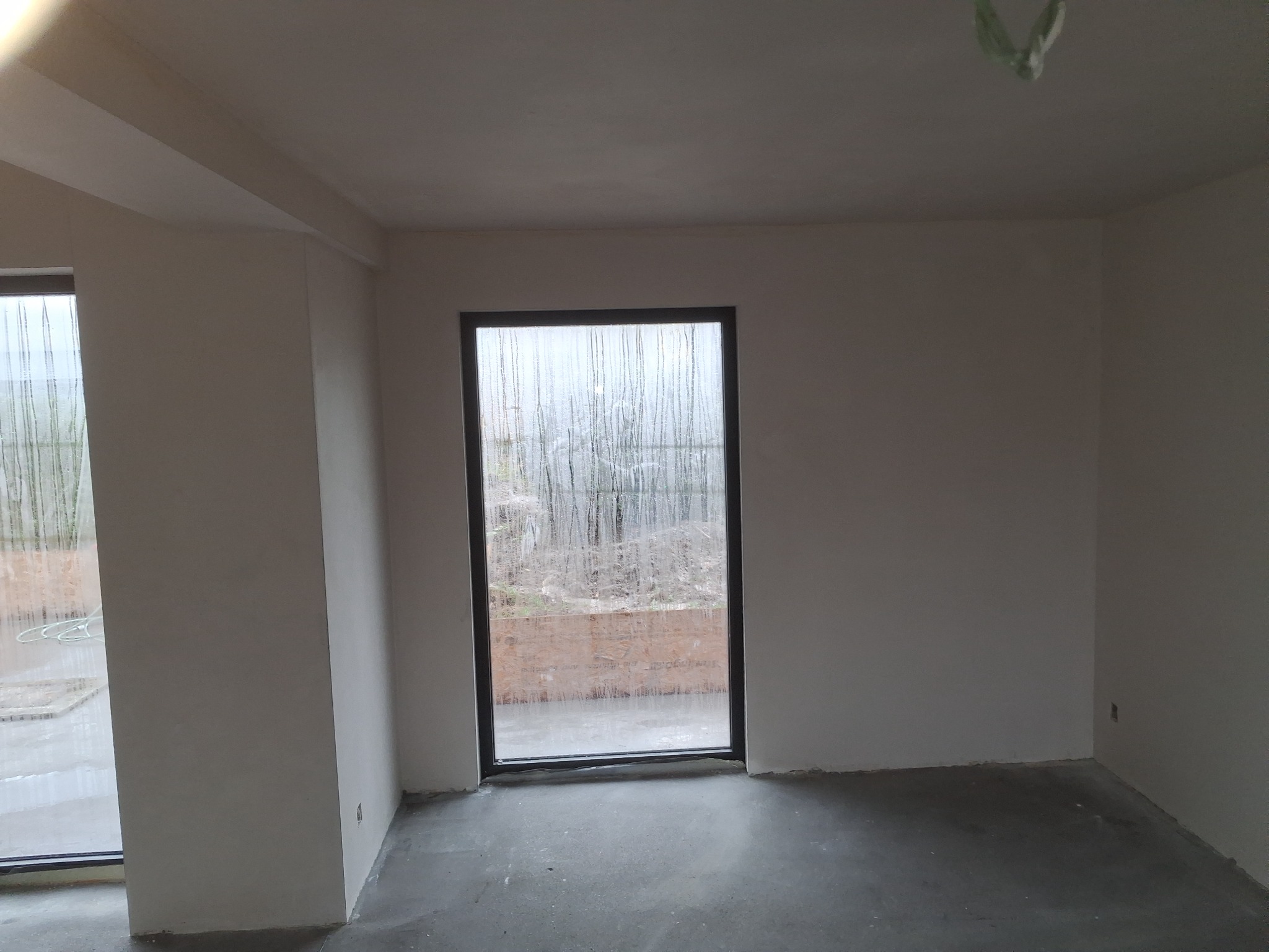 Wnętrze po gładziowaniu: jasne ściany, surowa podłoga, okno z widokiem na zewnątrz. Minimalistyczne wykończenie, gotowe do dalszych prac.