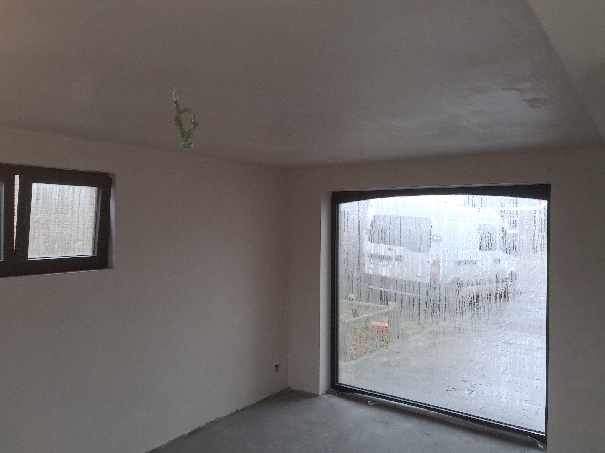 Wnętrze po tynkowaniu: gładkie ściany, okno z widokiem na zewnątrz, wystające przewody elektryczne. Świeżo położone tynki w jasnym pomieszczeniu.