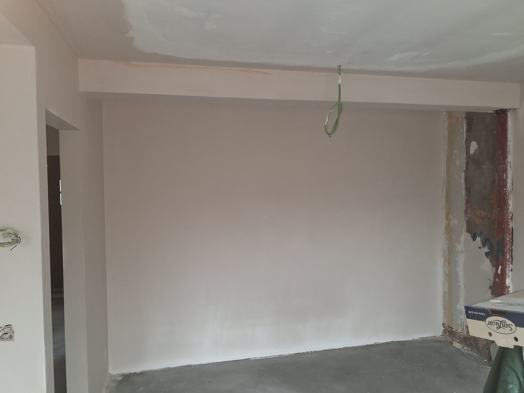 Wnętrze po gładziowaniu ścian, widoczne ślady po instalacji elektrycznej. Ściany w jasnym odcieniu, sufit biały. Fragment kolumny w stanie surowym. Podłoga szara, betonowa.