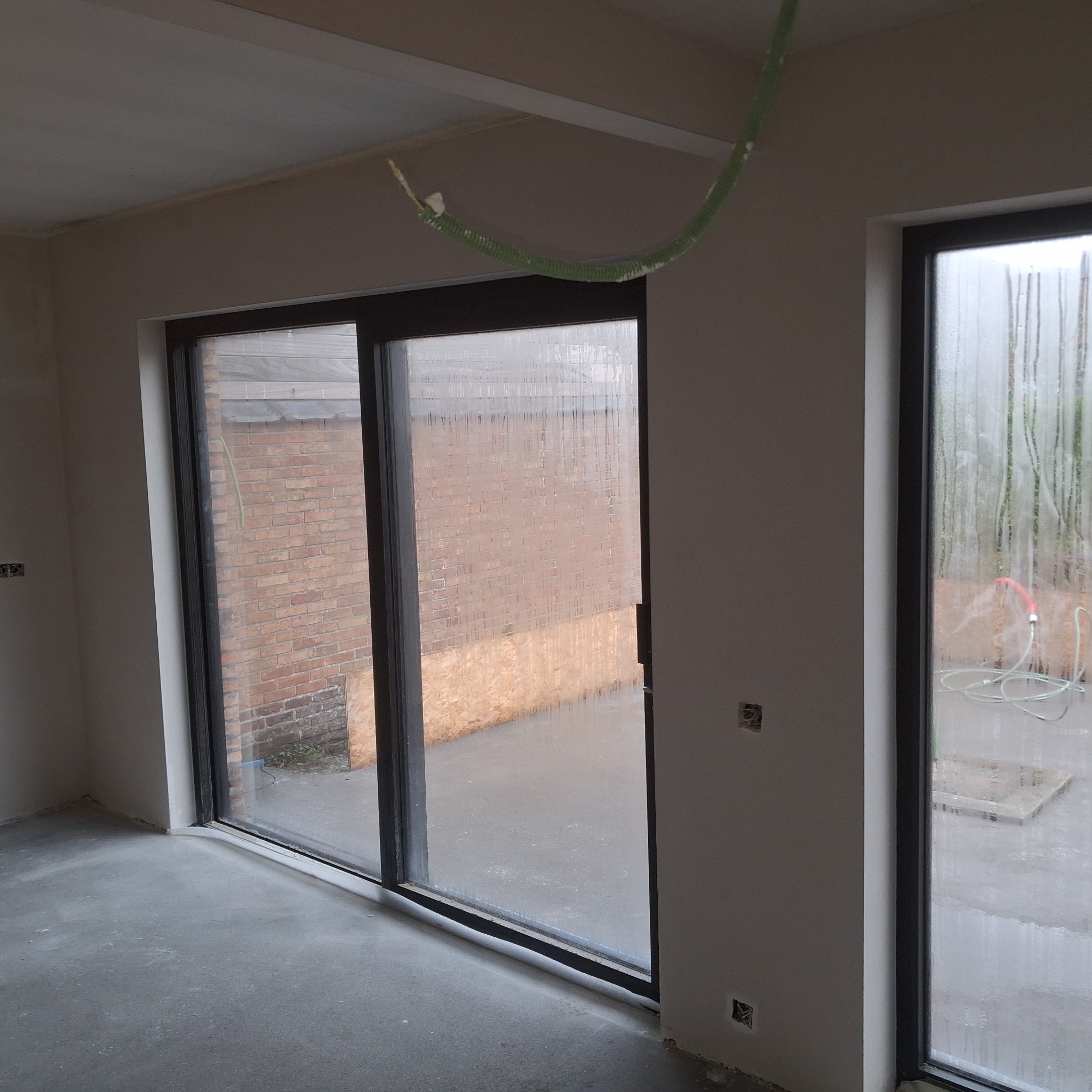 Surowe wnętrze z dużym oknem przesuwnym i widoczną instalacją elektryczną. Ściany przygotowane do dalszych prac wykończeniowych, surowy beton na podłodze.