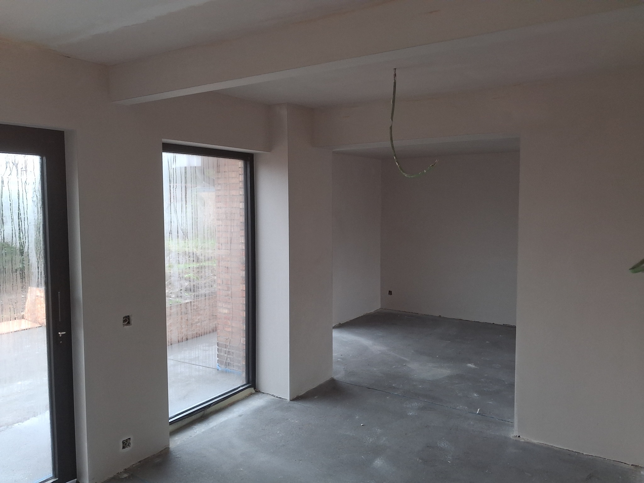 Wnętrze w stanie deweloperskim z oknami, świeżo otynkowanymi ścianami i surową podłogą. Widoczne wystające przewody elektryczne. Okna z widokiem na ceglany budynek.