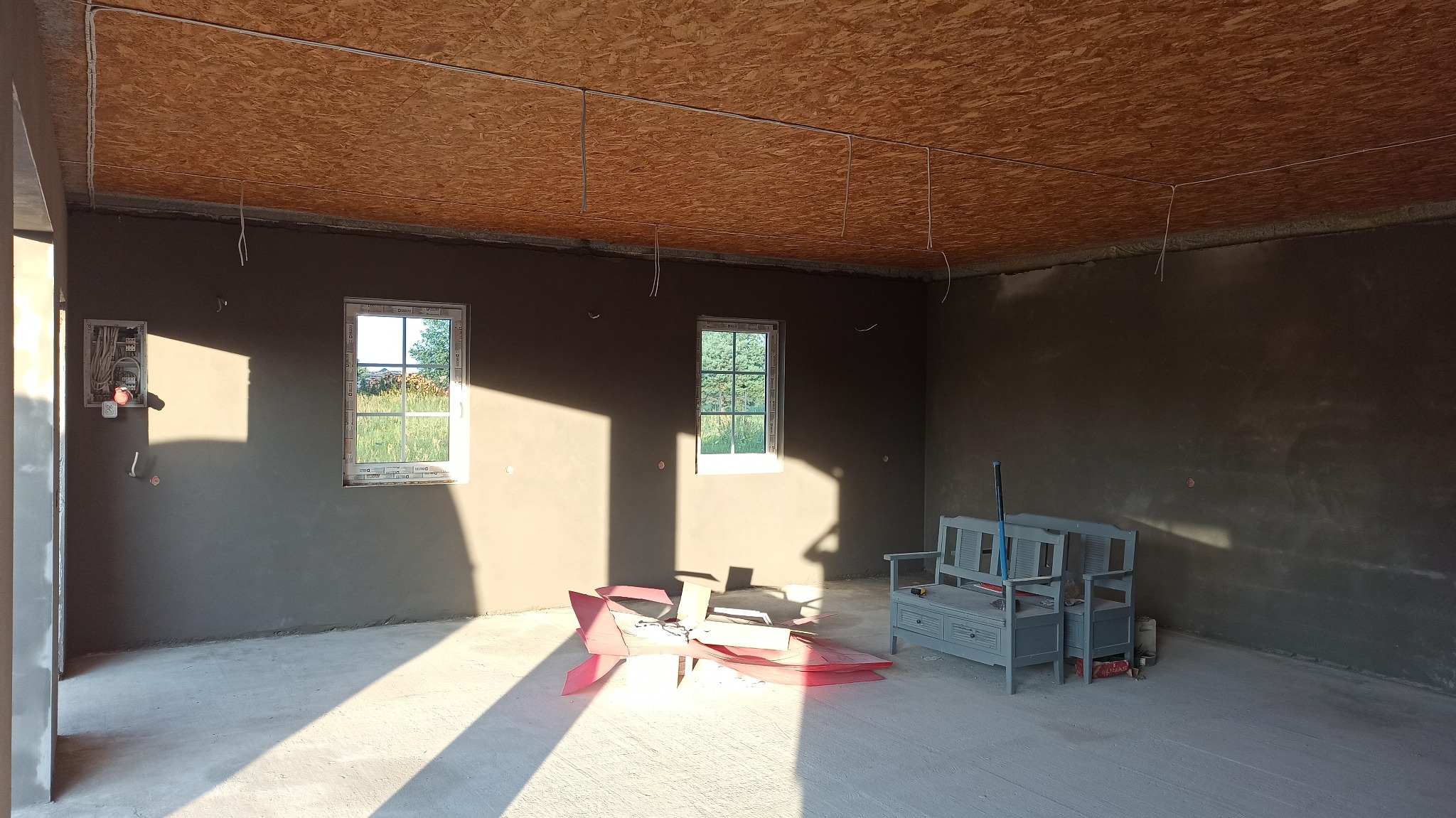 Surowe wnętrze z oknami po tynkowaniu, widoczne instalacje elektryczne pod sufitem z płyty OSB, resztki folii malarskiej na podłodze i ławka.