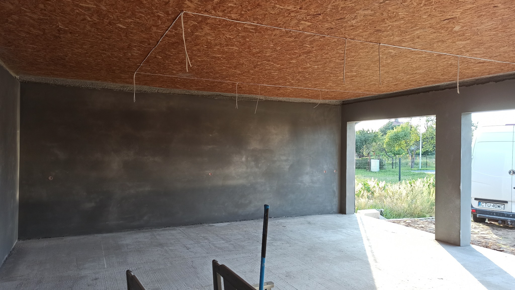 Surowe wnętrze w trakcie prac wykończeniowych: ściany pokryte świeżym tynkiem, widoczne przewody elektryczne pod sufitem z płyty OSB, narzędzie oparty o ścianę.