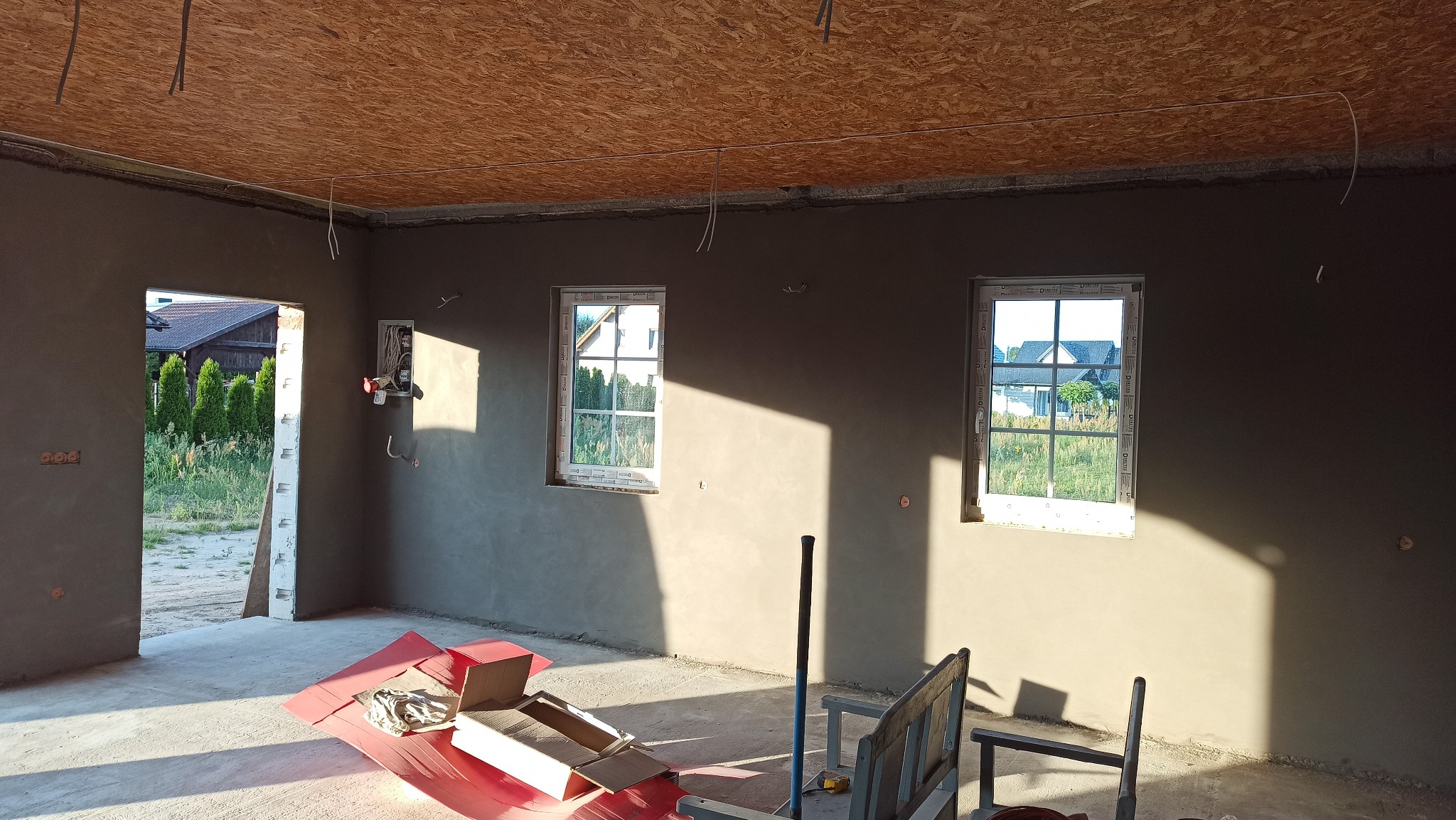 Wnętrze w stanie surowym zamkniętym: ściany pokryte świeżym tynkiem, dwa okna z widokiem na zieleń, sufit z płyt OSB, widoczne instalacje elektryczne.
