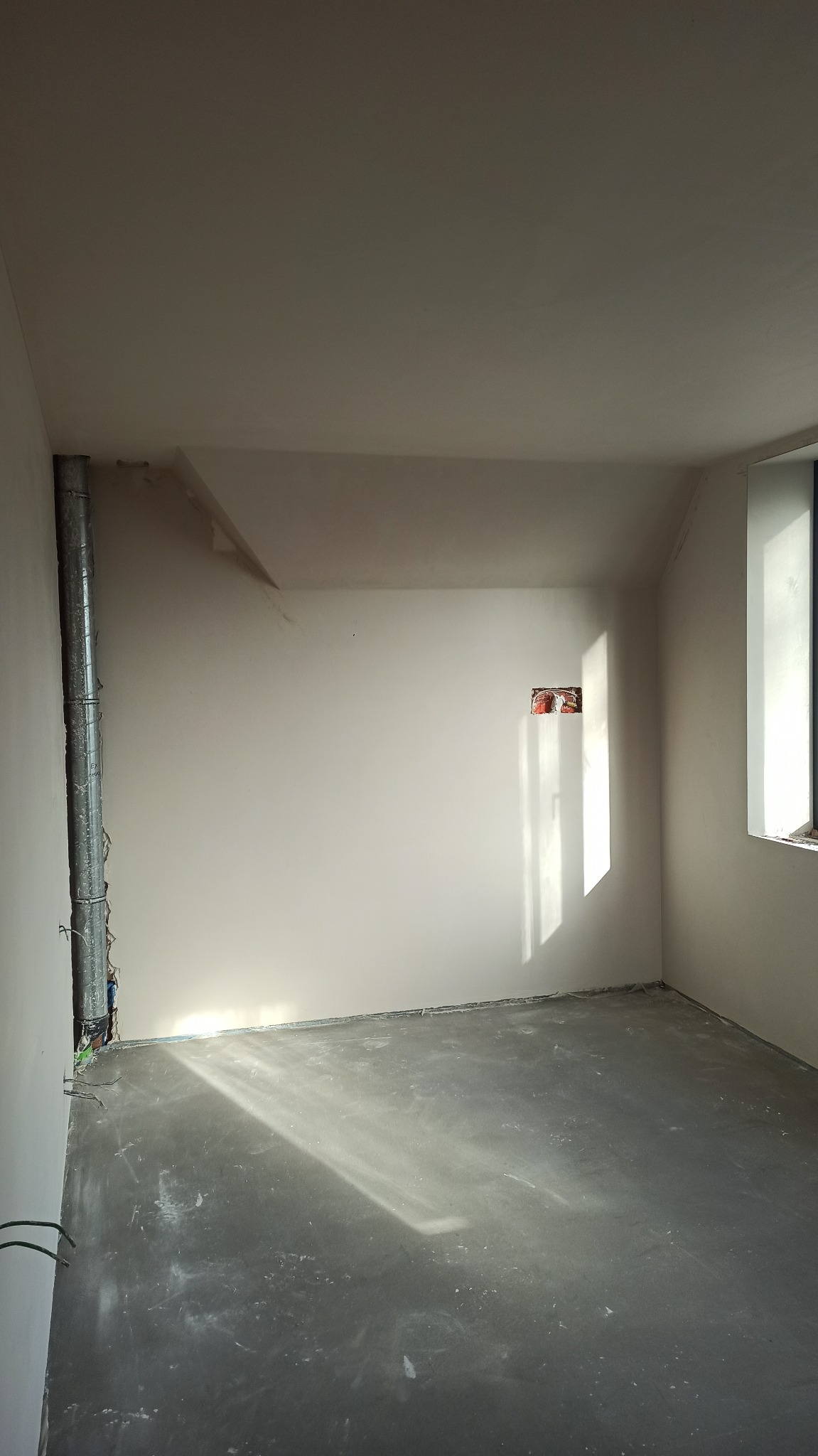 Pokój po tynkowaniu maszynowym: gładkie, białe ściany, szara posadzka, widoczne instalacje przy rurze wentylacyjnej, światło słoneczne z okna. Surowy stan wykończenia.