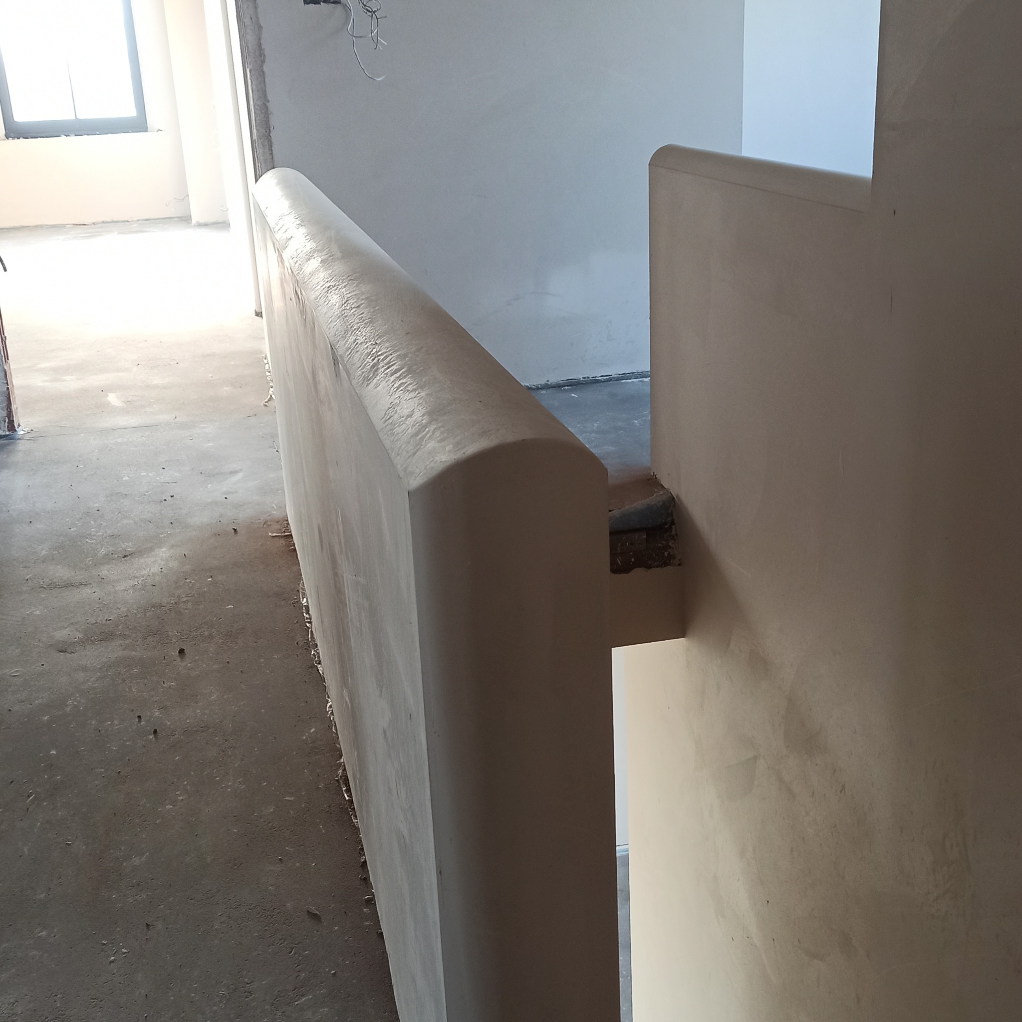 Świeżo otynkowana balustrada schodowa z zaokrąglonym wykończeniem, widoczna w surowym wnętrzu budynku w trakcie prac wykończeniowych.