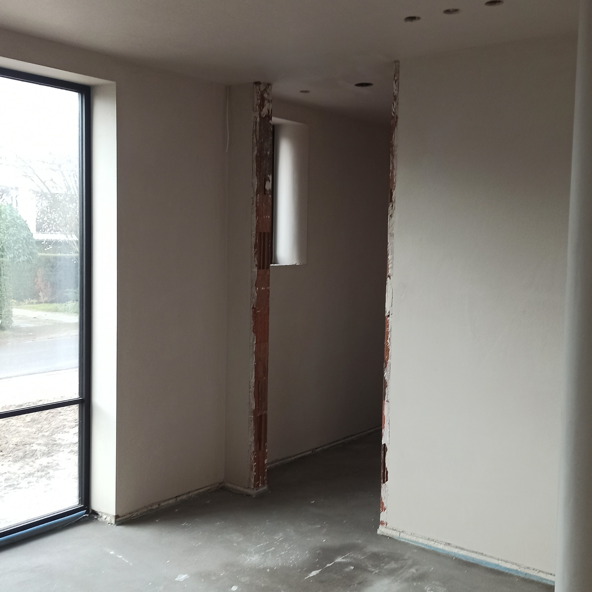 Wnętrze w trakcie prac wykończeniowych: gładkie, jasne ściany po tynkowaniu, surowa podłoga, widoczne fragmenty cegieł przy otworach drzwiowych, duże okno.