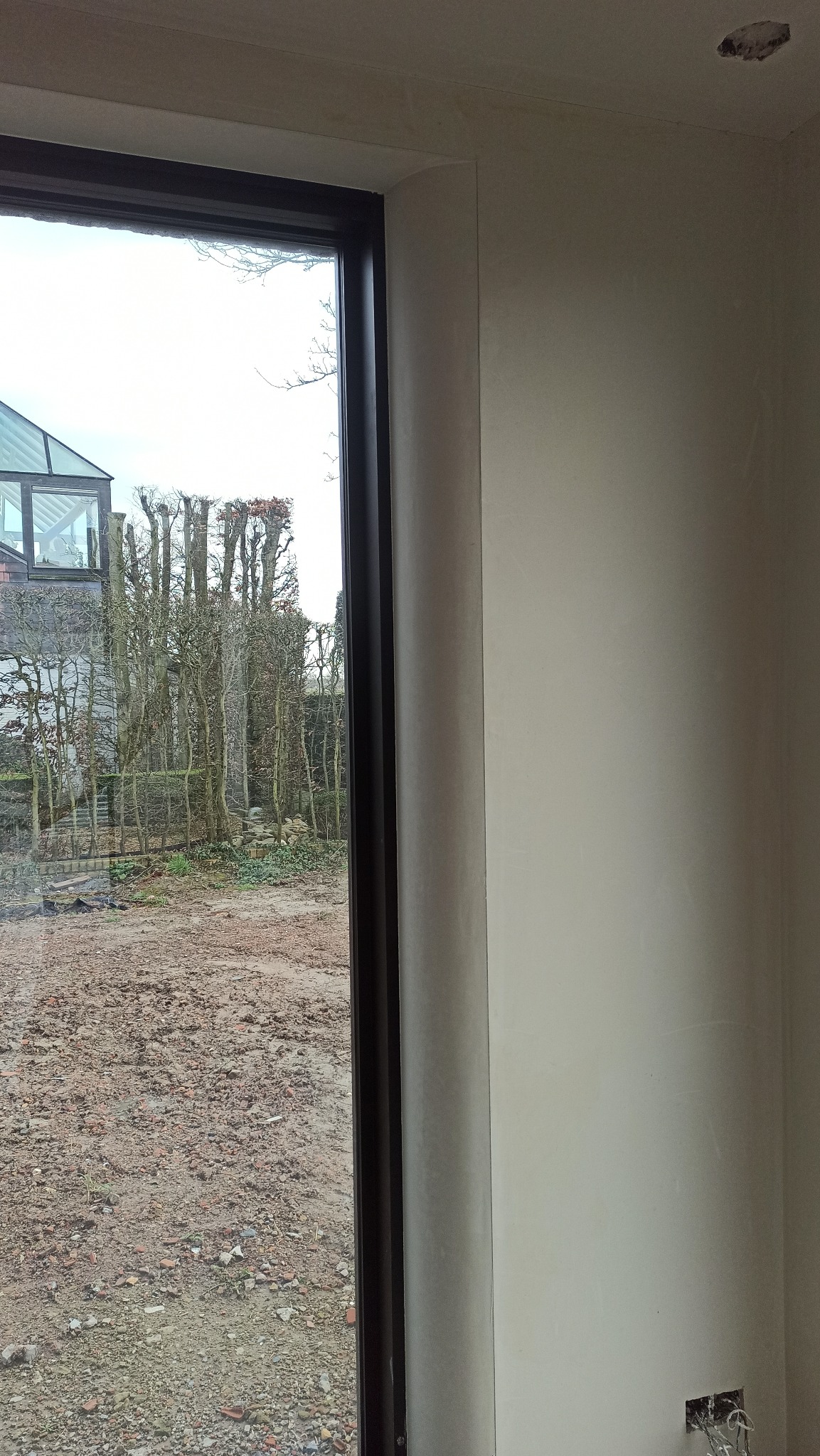 Gładka, jasna ściana po tynkowaniu, z widocznym oknem z czarną ramą i widokiem na ogród. W dolnym rogu widoczne gniazdko elektryczne.