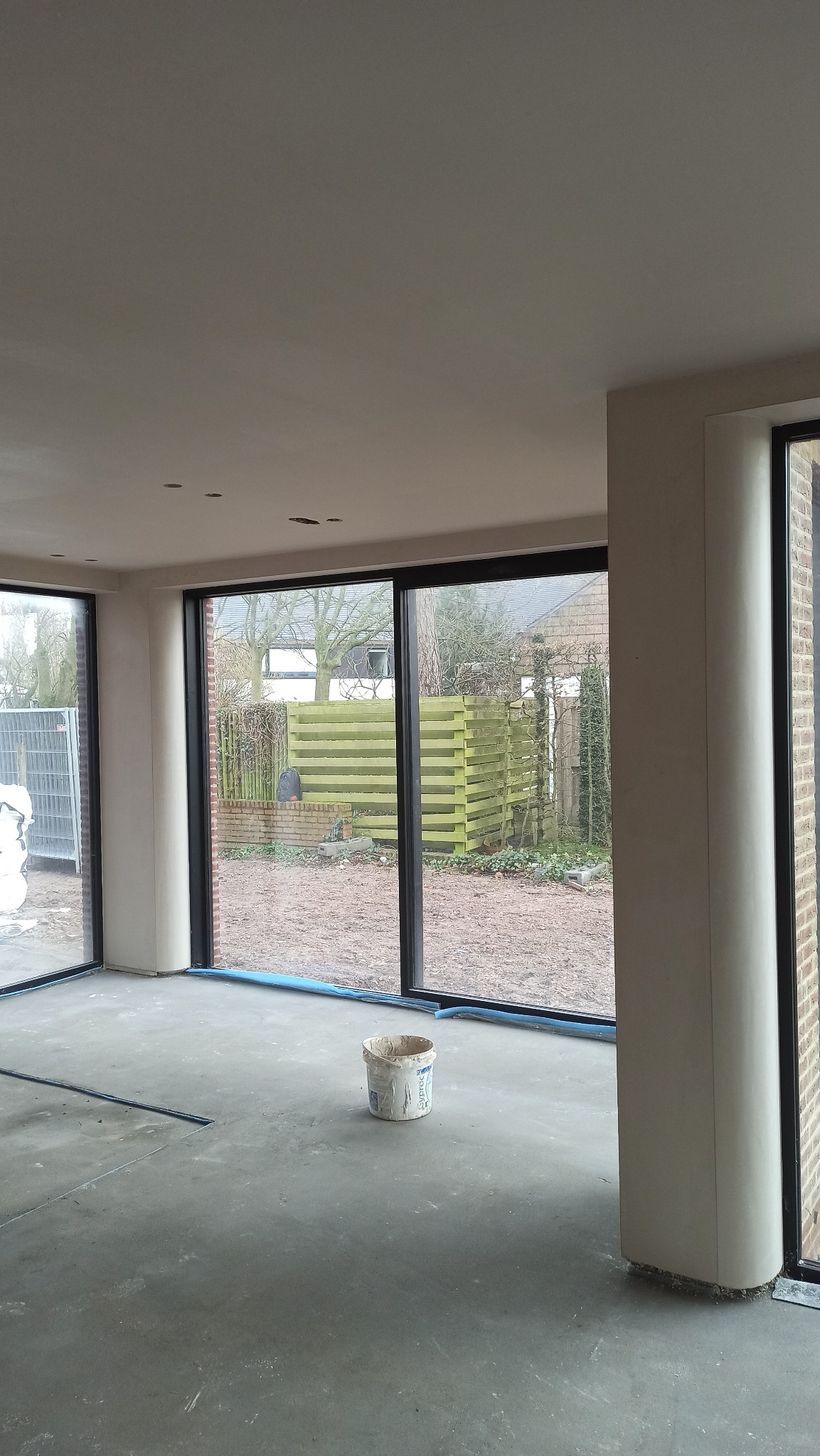 Wnętrze po tynkowaniu maszynowym: gładkie ściany, duże okna z widokiem na zewnątrz, wiadro po farbie na szarej posadzce. Widoczne ślady po pracach wykończeniowych.