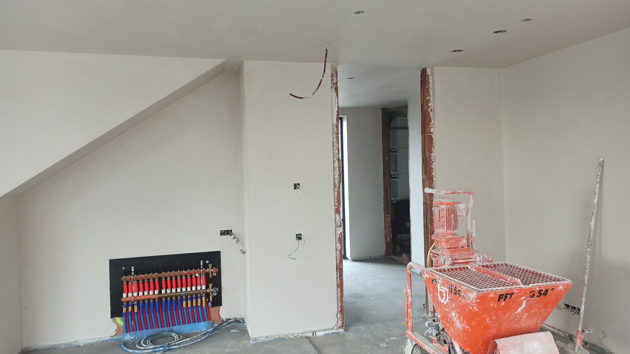 Wnętrze w trakcie prac wykończeniowych: świeżo otynkowane ściany, widoczna instalacja grzewcza podłogowa i maszyna do tynkowania PFT G54. Surowe wykończenie, gotowe do dalszych prac.