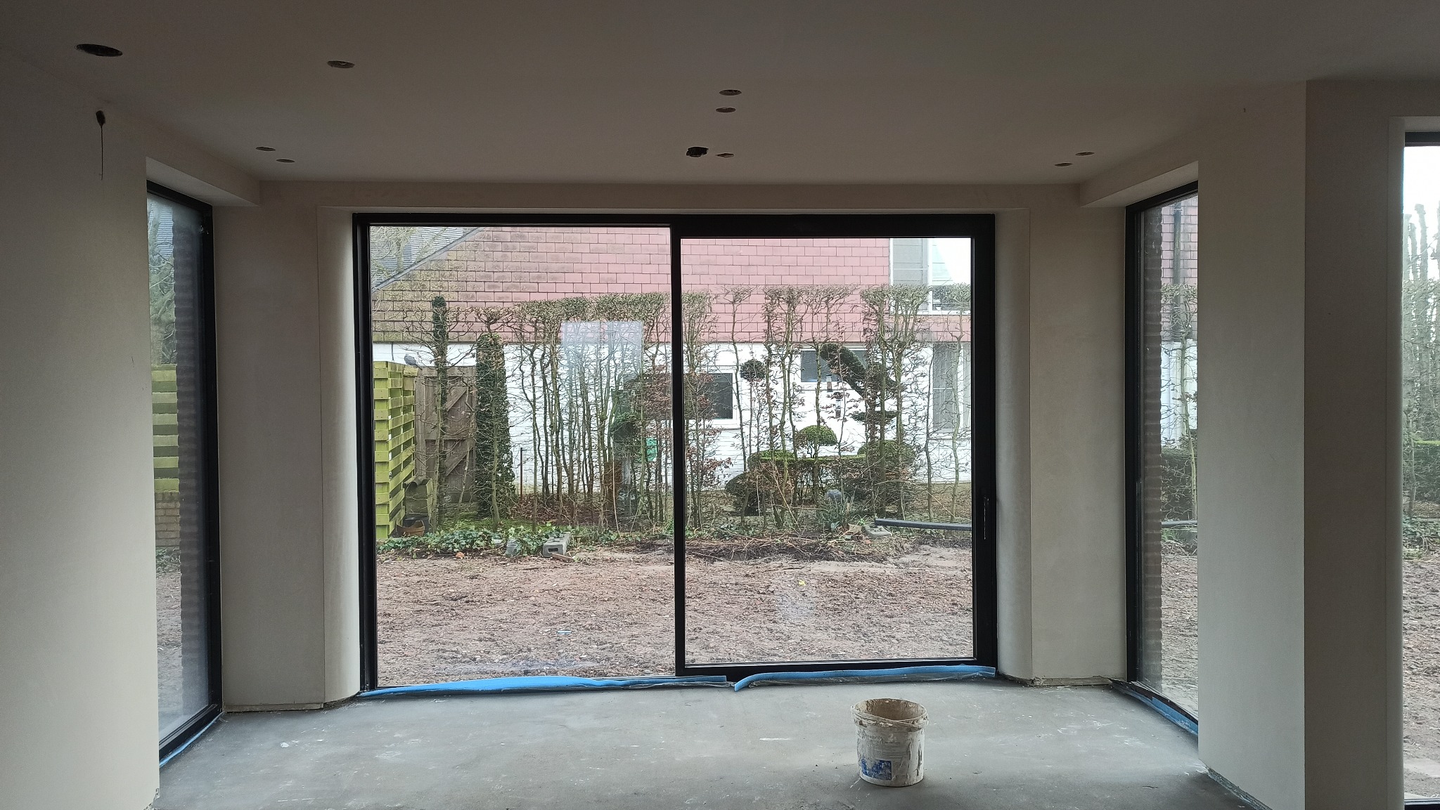 Wnętrze w trakcie prac wykończeniowych: ściany po gładziowaniu, zabezpieczona podłoga, duże okno przesuwne z widokiem na ogród. Widoczne niedokończone punkty oświetleniowe.