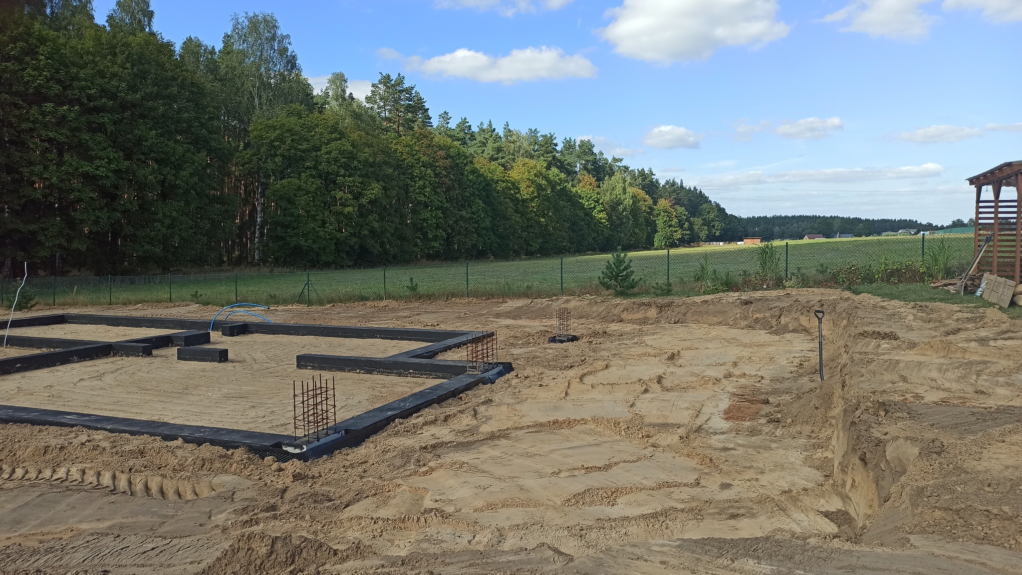 Przygotowany teren pod fundamenty domu jednorodzinnego. Widoczne zbrojenia, czarna izolacja, wykopane rowy i piasek. W tle las i błękitne niebo.