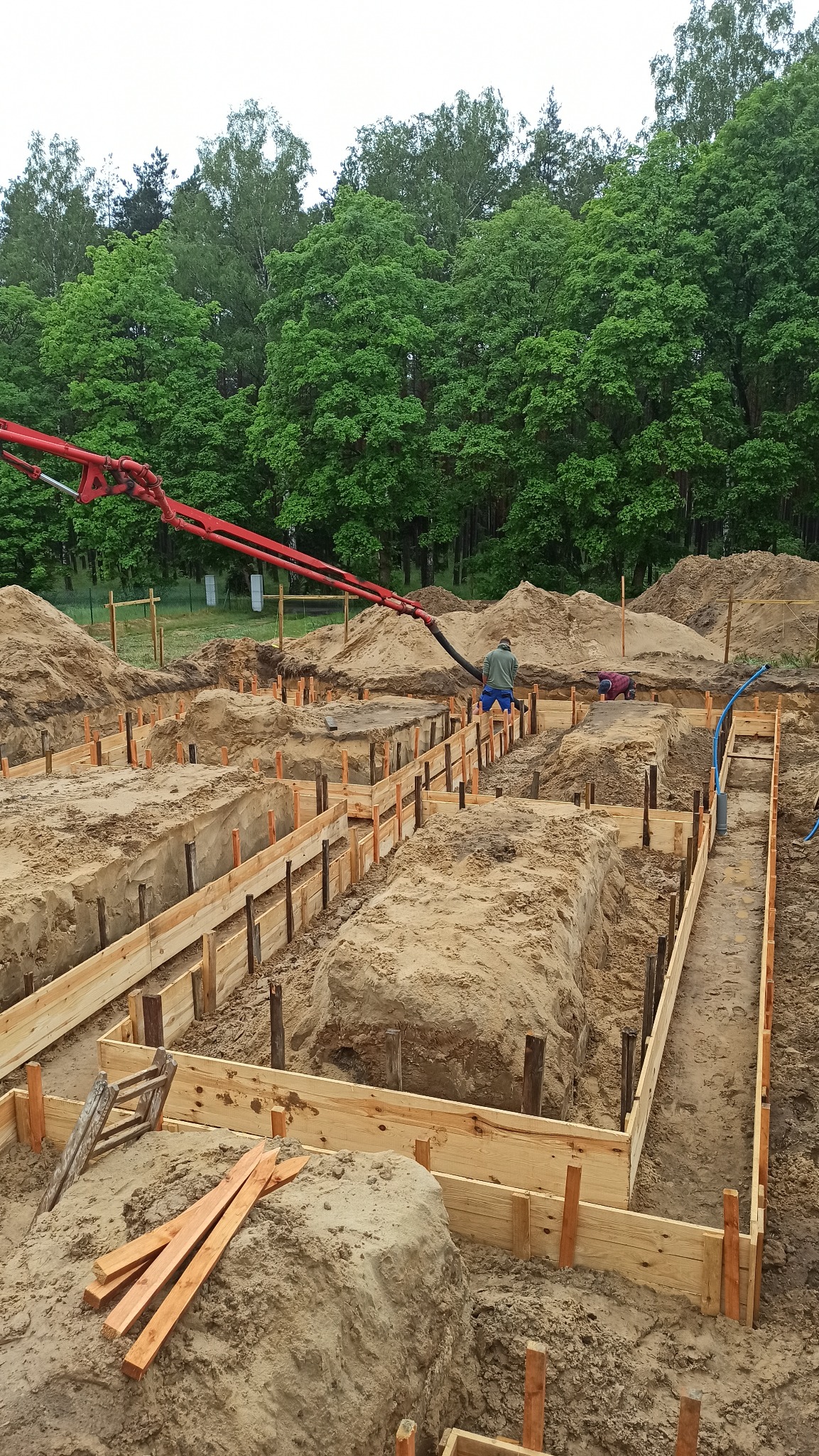Wykopane fundamenty z drewnianym szalunkiem, przygotowane do zalewania betonem. W tle widoczna pompa do betonu i zalesiony teren.