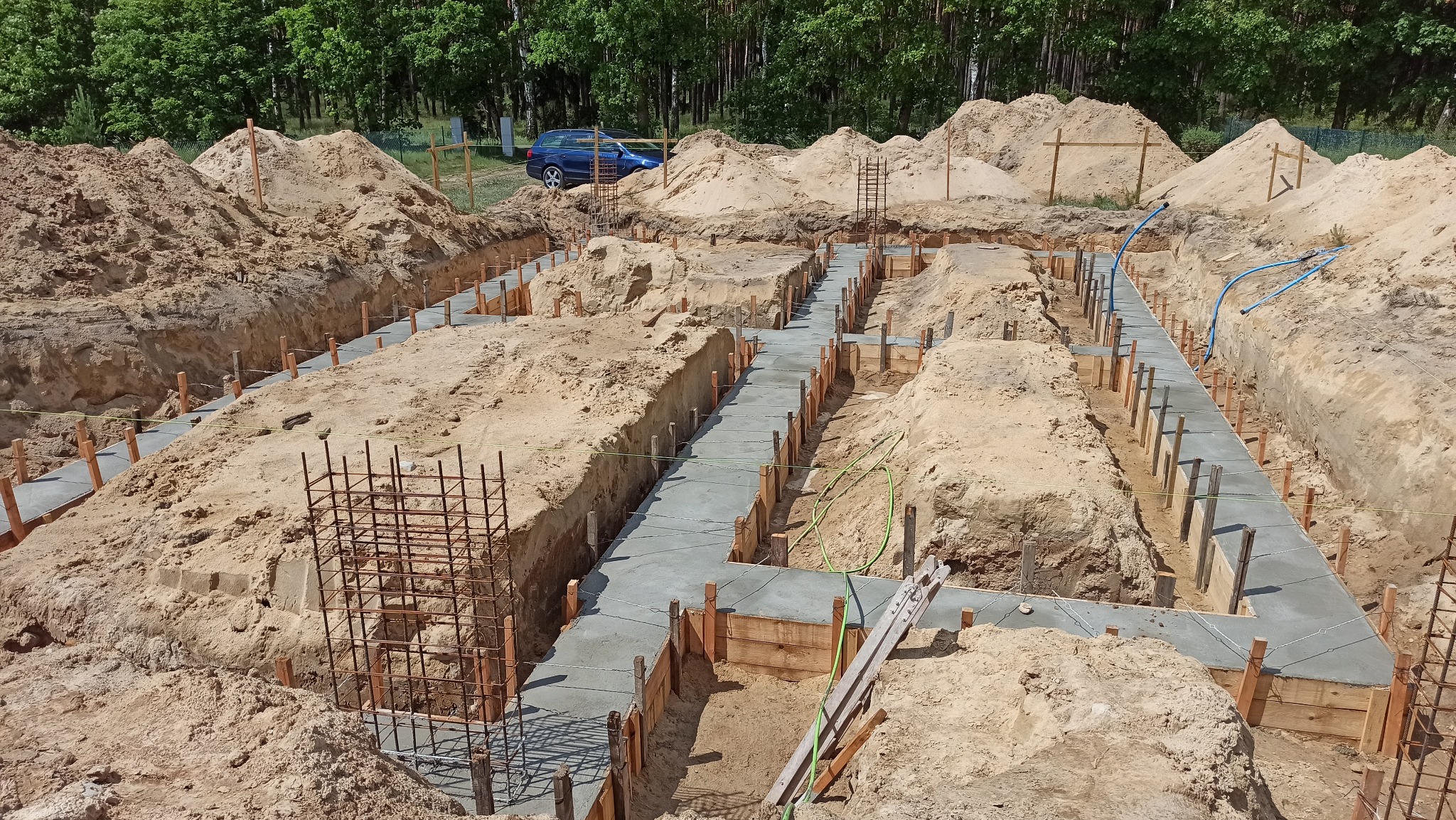 Zalane betonem fundamenty w wykopie, z widocznym zbrojeniem i deskowaniem. W tle hałdy piasku i fragment lasu. Ujęcie z góry, w słoneczny dzień.
