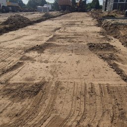 Usługi transportowe Robert Szybowski - Przygotowany teren pod budowę z widocznymi śladami maszyn, w tle ciężarówka, koparka i budynek gospodarczy. Błękitne niebo z chmurami.