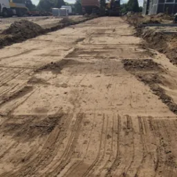 Przygotowany teren pod budowę z widocznymi śladami maszyn, w tle ciężarówka, koparka i budynek gospodarczy. Błękitne niebo z chmurami.