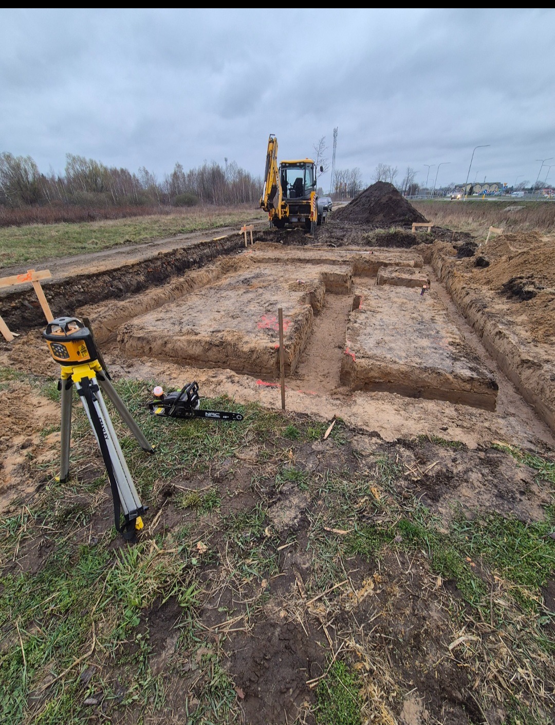 Wykop pod fundamenty z wytyczonym obrysem, widoczny sprzęt pomiarowy i koparka w tle. Teren przygotowany pod budowę w pochmurny dzień.