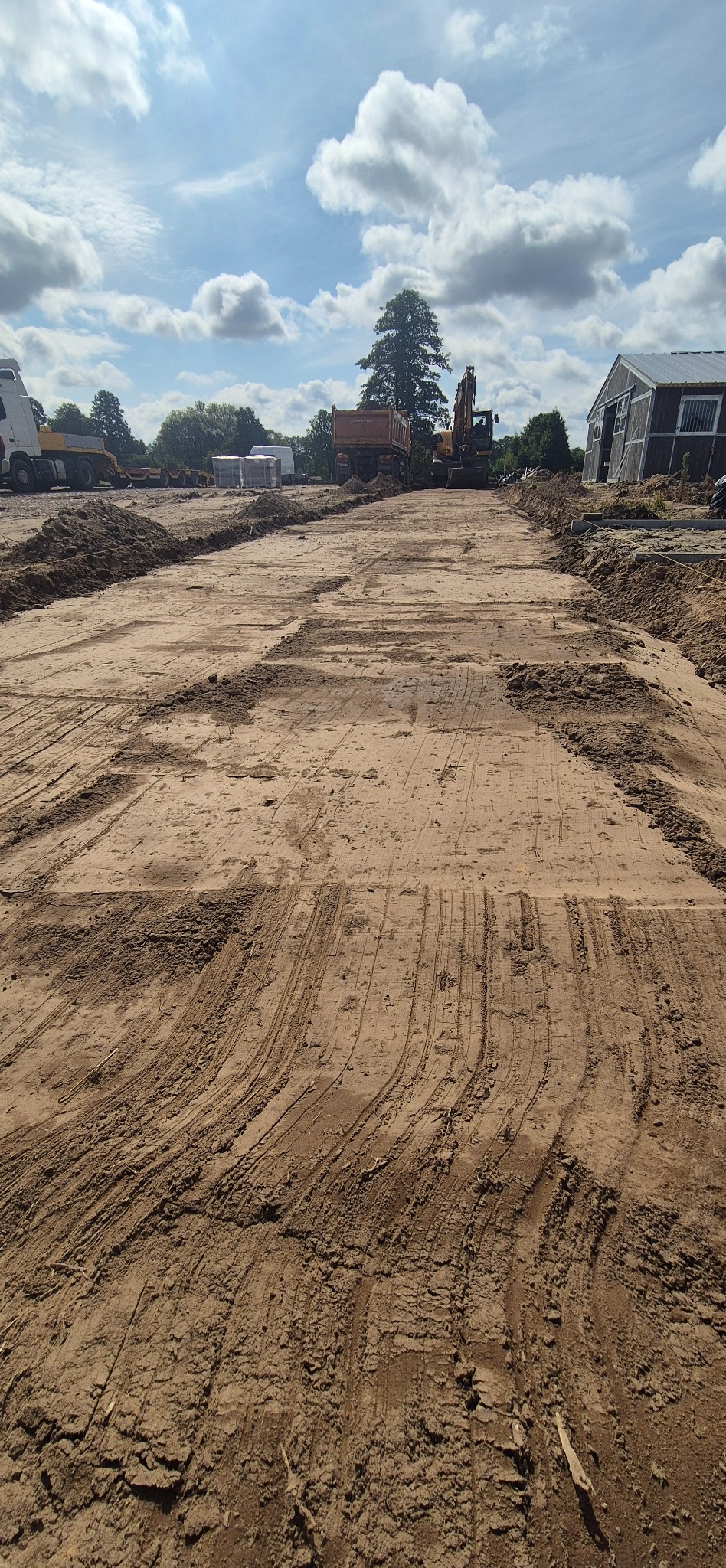 Przygotowany teren pod budowę z widocznymi śladami maszyn, w tle ciężarówka, koparka i budynek gospodarczy. Błękitne niebo z chmurami.