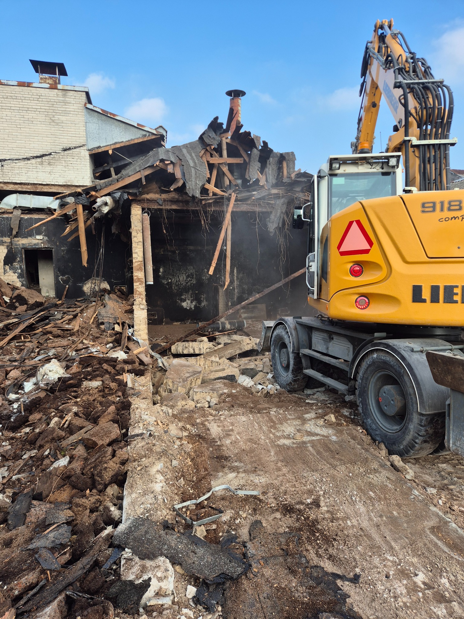 Żółta koparka Liebherr 918 na tle częściowo zburzonego, czarnego budynku z drewnianymi elementami. Gruz i ziemia na pierwszym planie, błękitne niebo w tle.