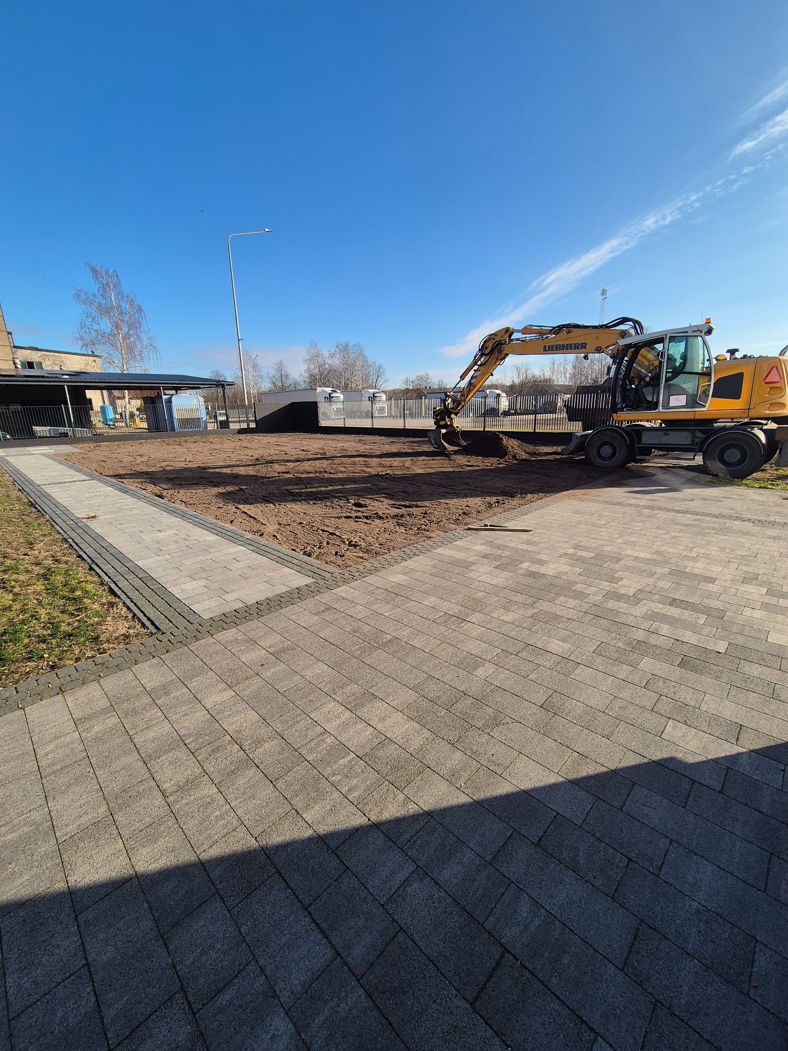 Żółta koparka Liebherr podczas prac ziemnych na działce z chodnikiem z kostki brukowej. Błękitne niebo w tle. Przygotowanie terenu pod inwestycję.