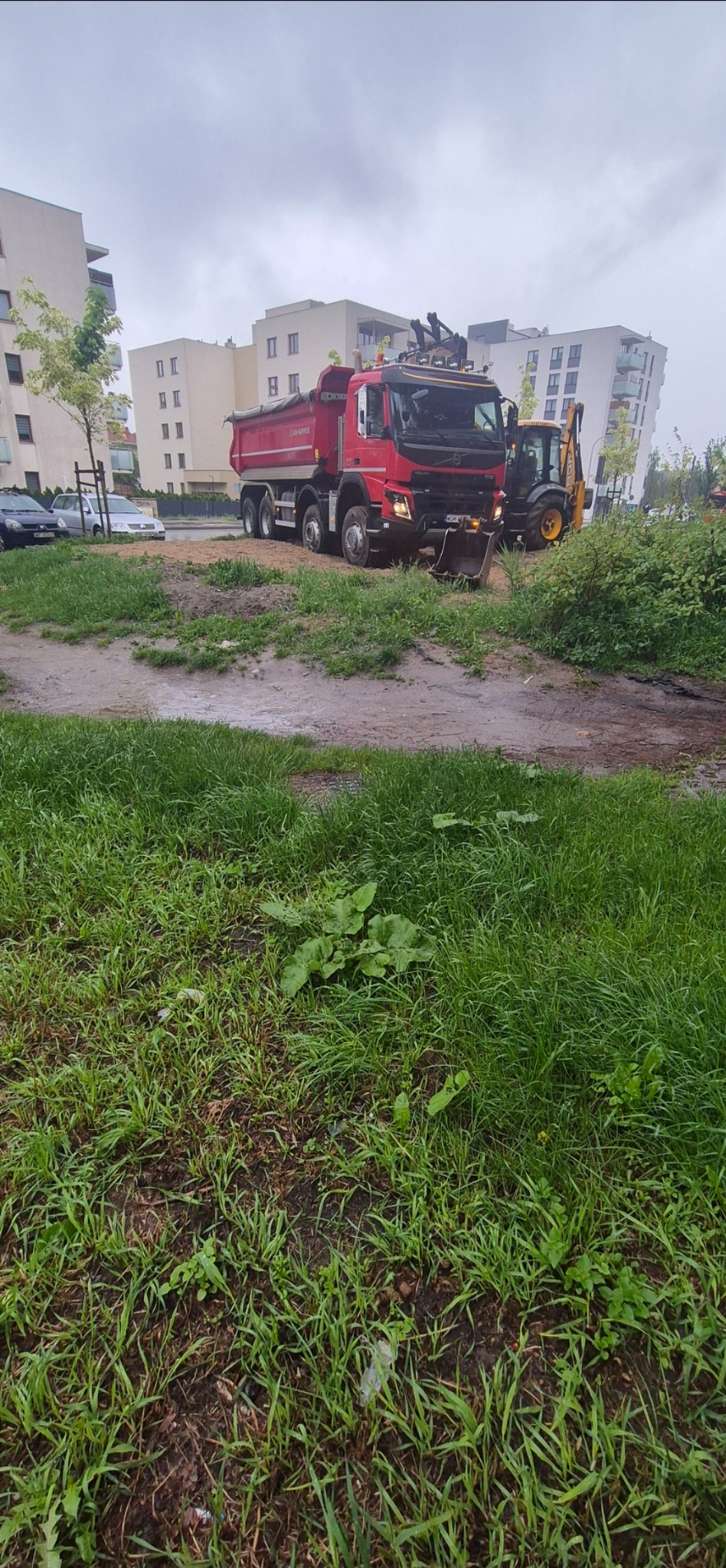 Czerwona wywrotka i koparka na terenie budowy, w tle bloki mieszkalne. Sprzęt gotowy do pracy, teren przygotowany pod inwestycję.