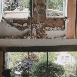 GENERAL-BUD SPÓŁKA Z OGRANICZONĄ ODPOWIEDZIALNOŚCIĄ - Widok przez otwór okienny po wyburzeniu ściany z cegły. Na dole pracownik w stroju roboczym. Za oknem zieleń drzew. Prace zabezpieczone podporami.